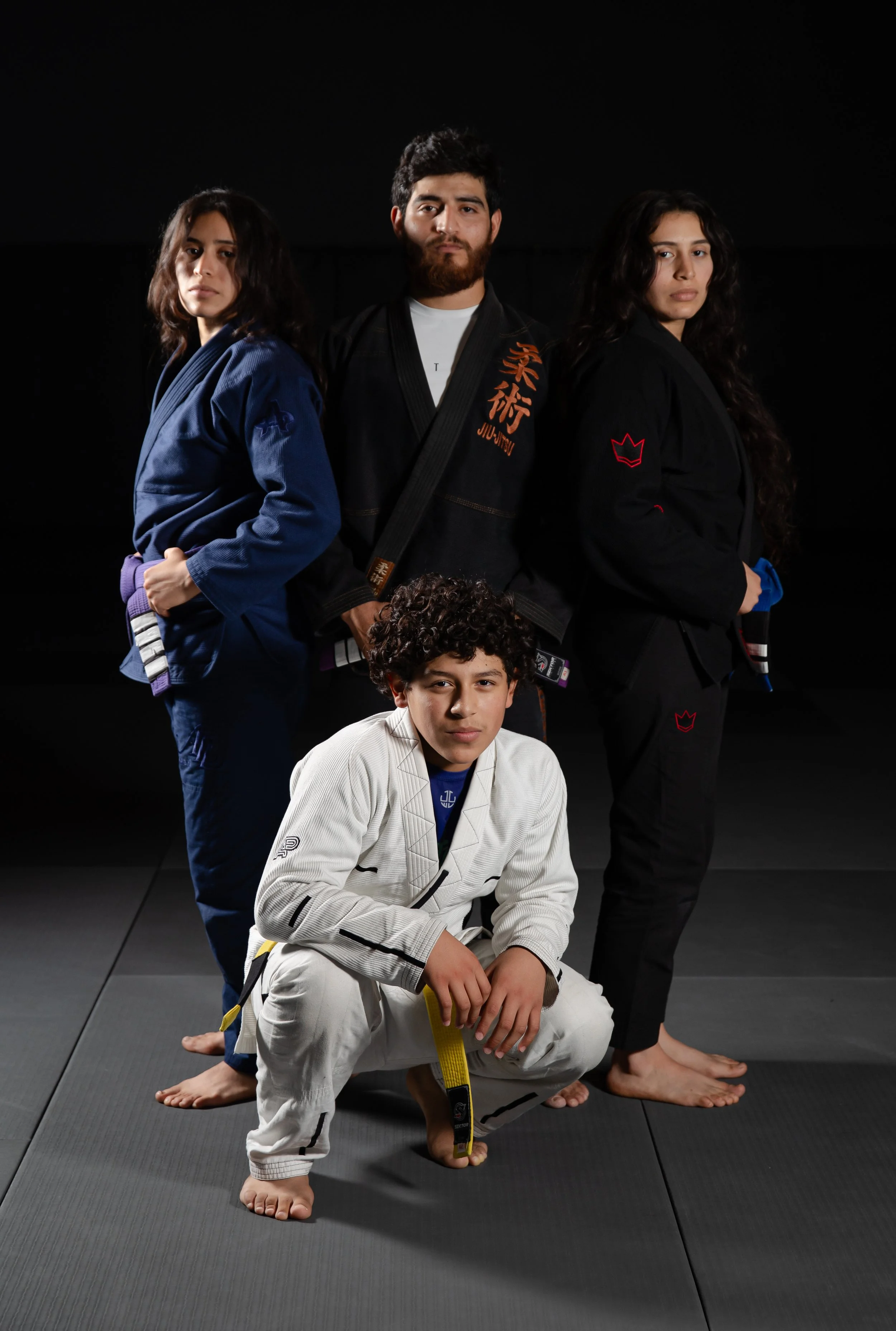 Group of four martial artists in Brazilian Jiu-Jitsu gis posing on a black mat in a dark training facility.