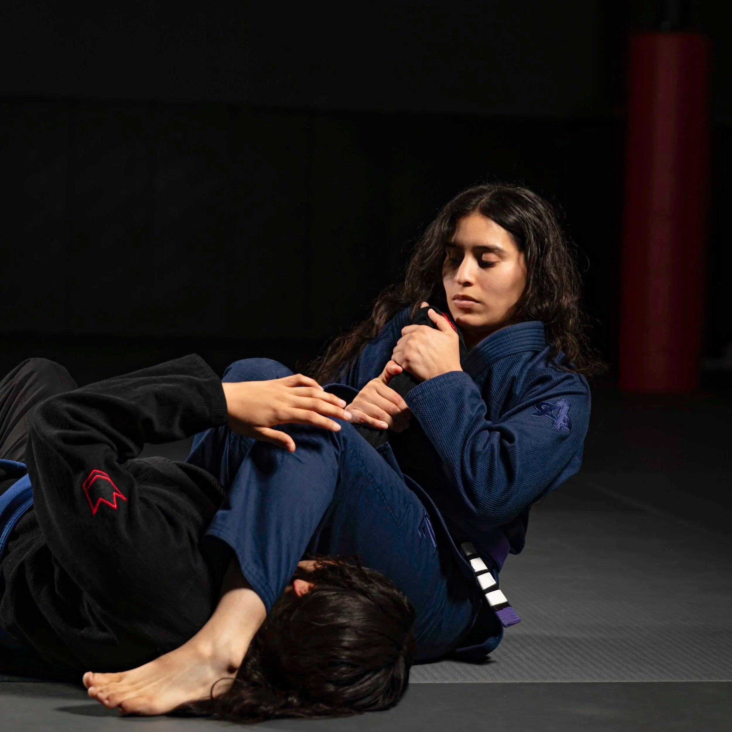 A woman with long curly hair lying on the floor with her eyes closed, holding her temples, possibly practicing meditation or relaxation. She is wearing a dark blue hoodie and black pants, with a dark background.