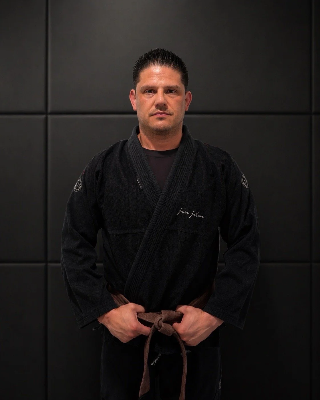 A man standing in a martial arts uniform in front of a black wall, posing with his hands at his waist.