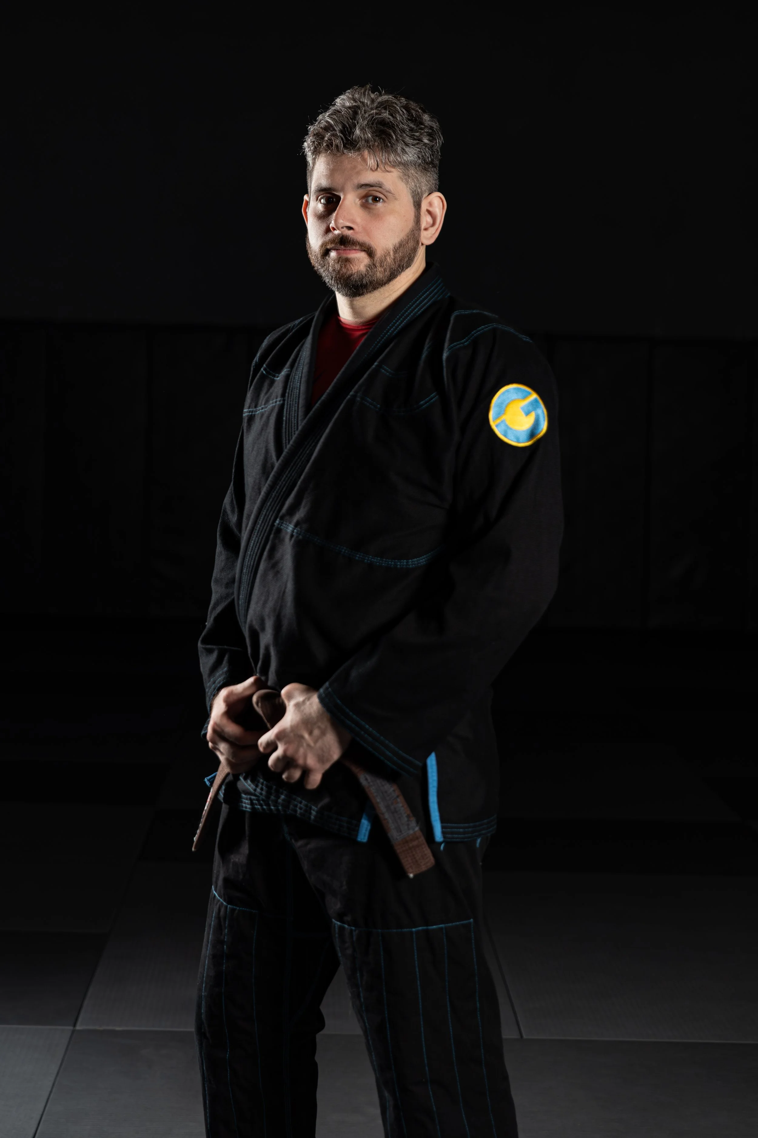 A man wearing a Brazilian Jiu-Jitsu gi with a patch on his left sleeve, standing in a martial arts training studio with a dark background and black mats.