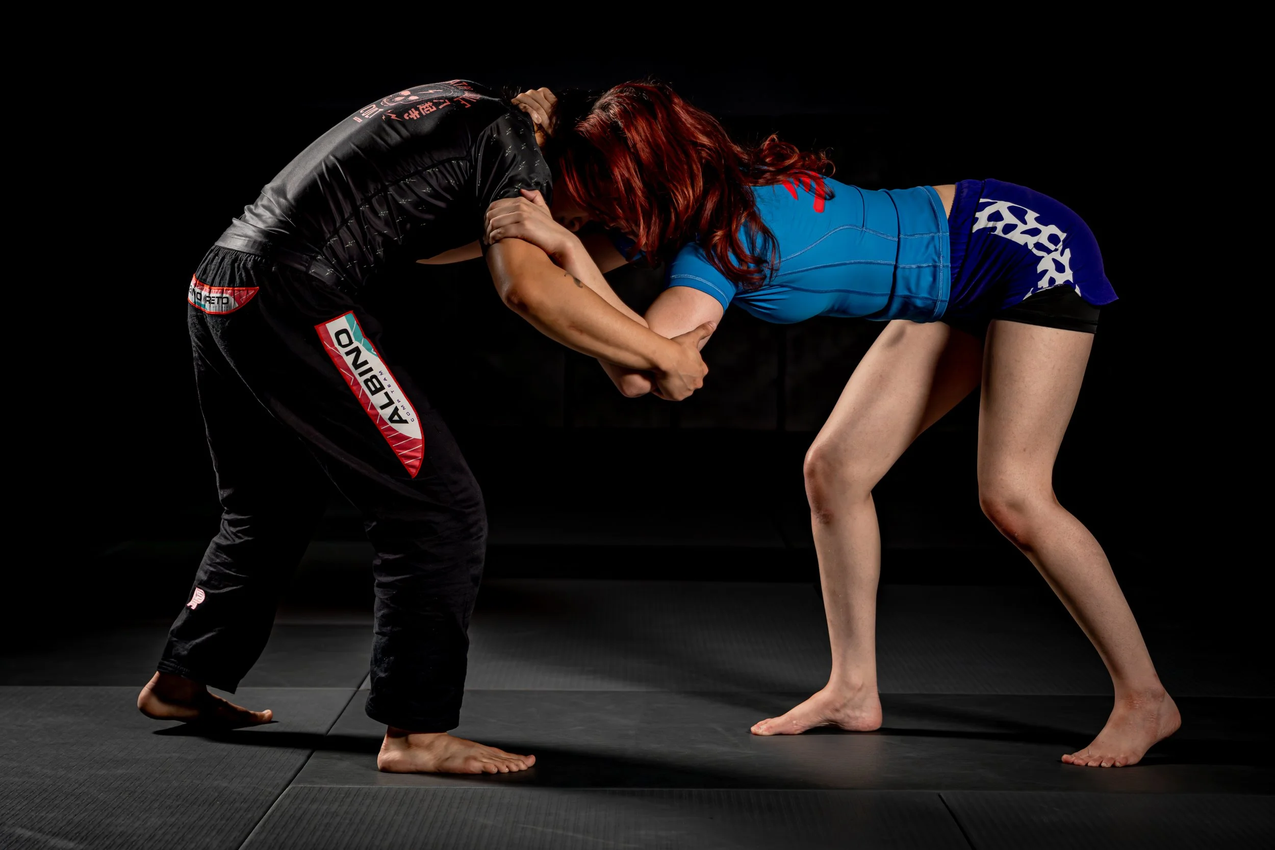 Two women practicing Brazilian Jiu-Jitsu on black mats in a dark training room, engaged in a grappling move.