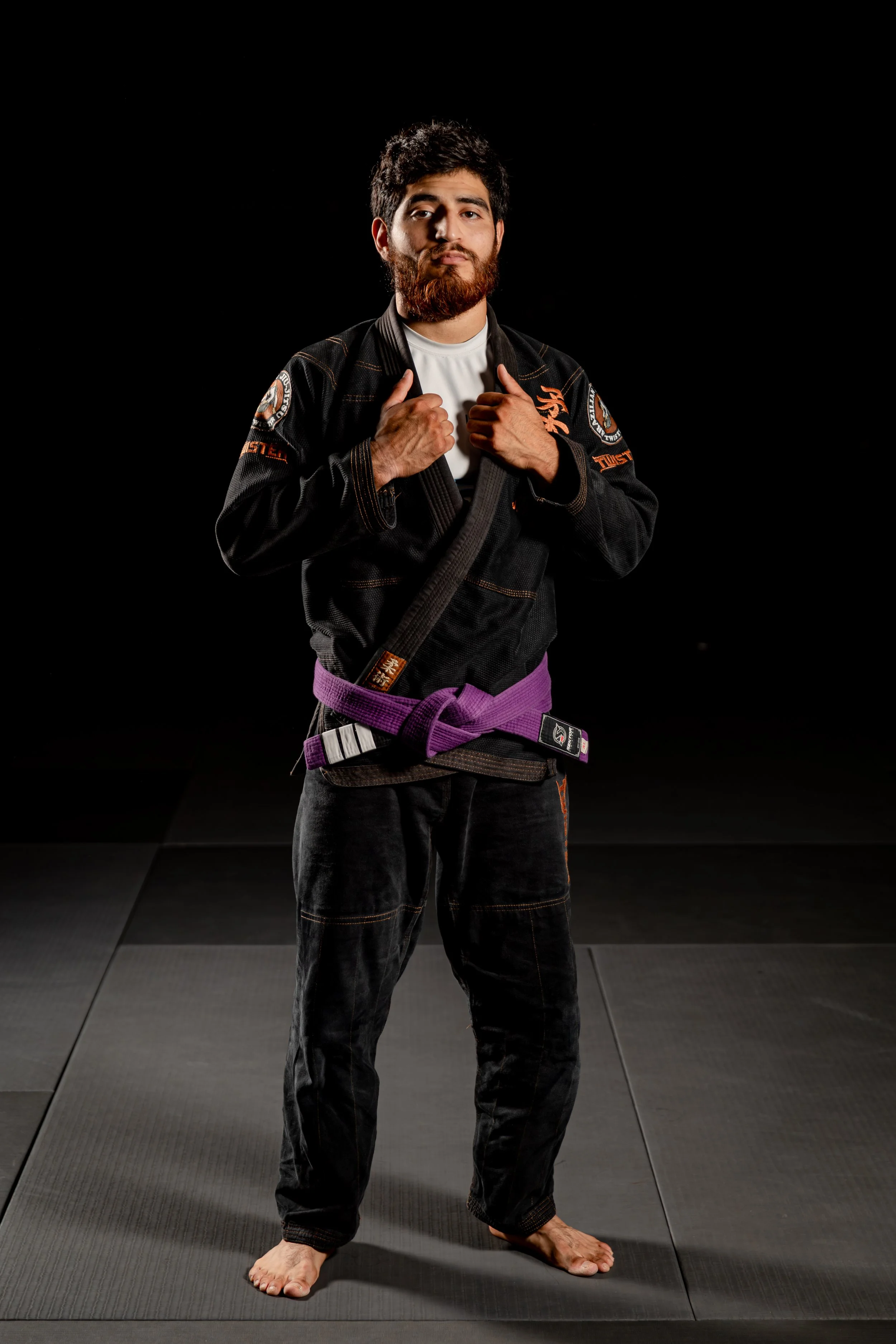 A man wearing a Brazilian jiu-jitsu gi standing barefoot on a dark mat, holding his collar with both hands, on a black background.
