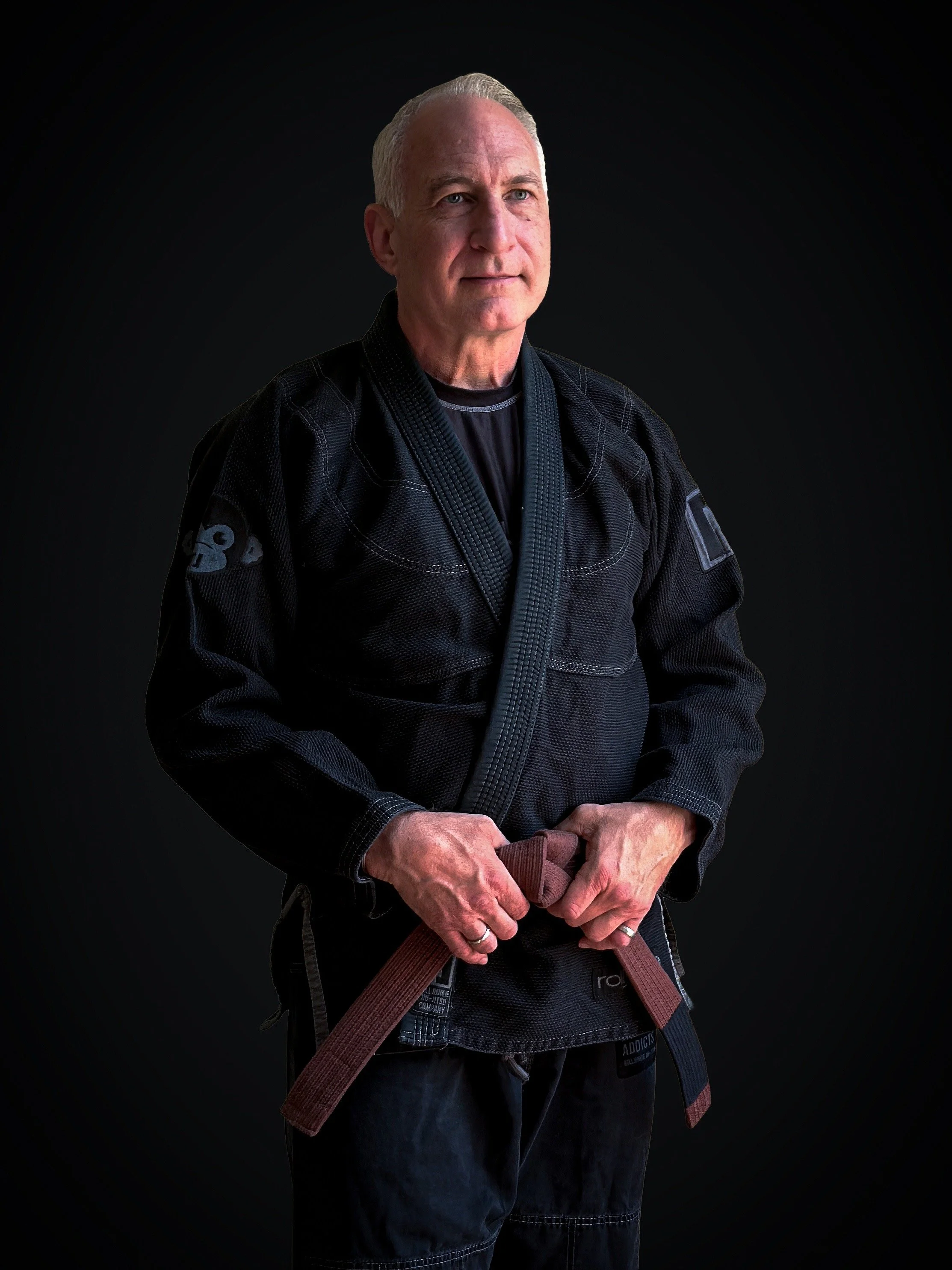 An older man with gray hair tied back, wearing a black martial arts uniform with patches and a brown belt, standing against a black background.
