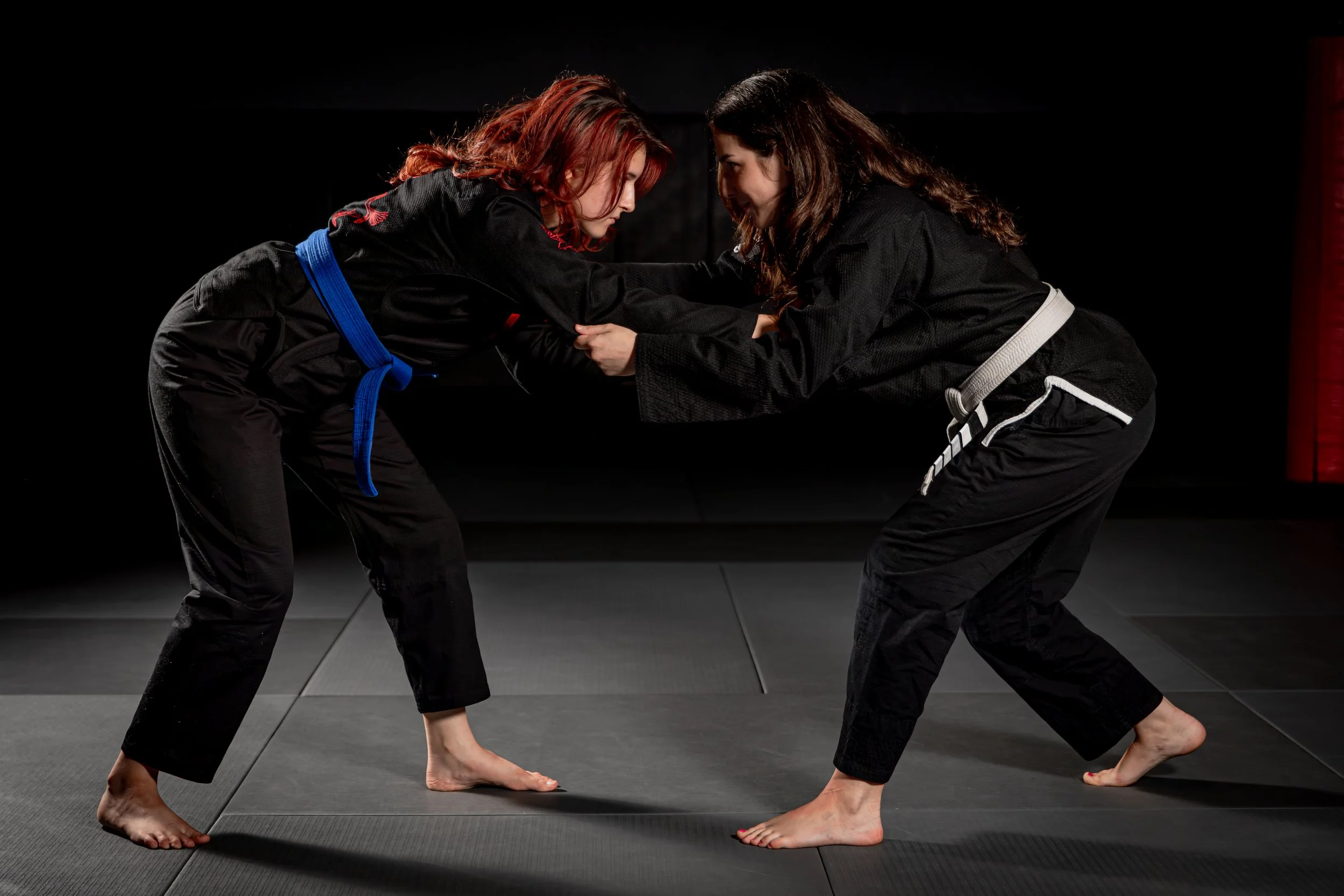 Two women practicing Brazilian Jiu-Jitsu on a black mat, engaging in a grappling hold, wearing black gis, in a dark indoor environment.