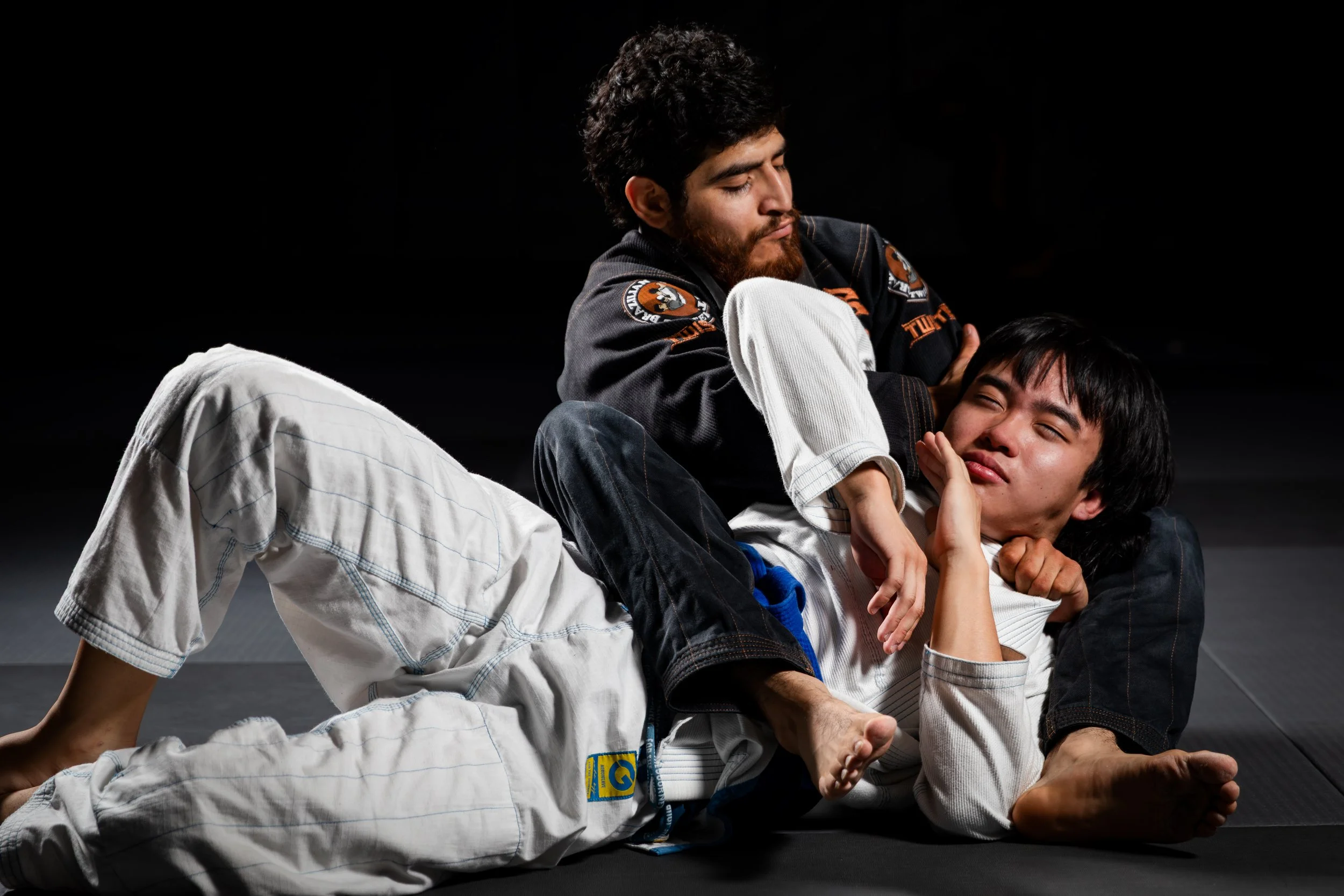 Two men practicing Brazilian Jiu-Jitsu on a black mat with a dark background. One man is wearing a white gi, and the other is wearing a black gi. They are engaged in a grappling position, with one man pinning the other.