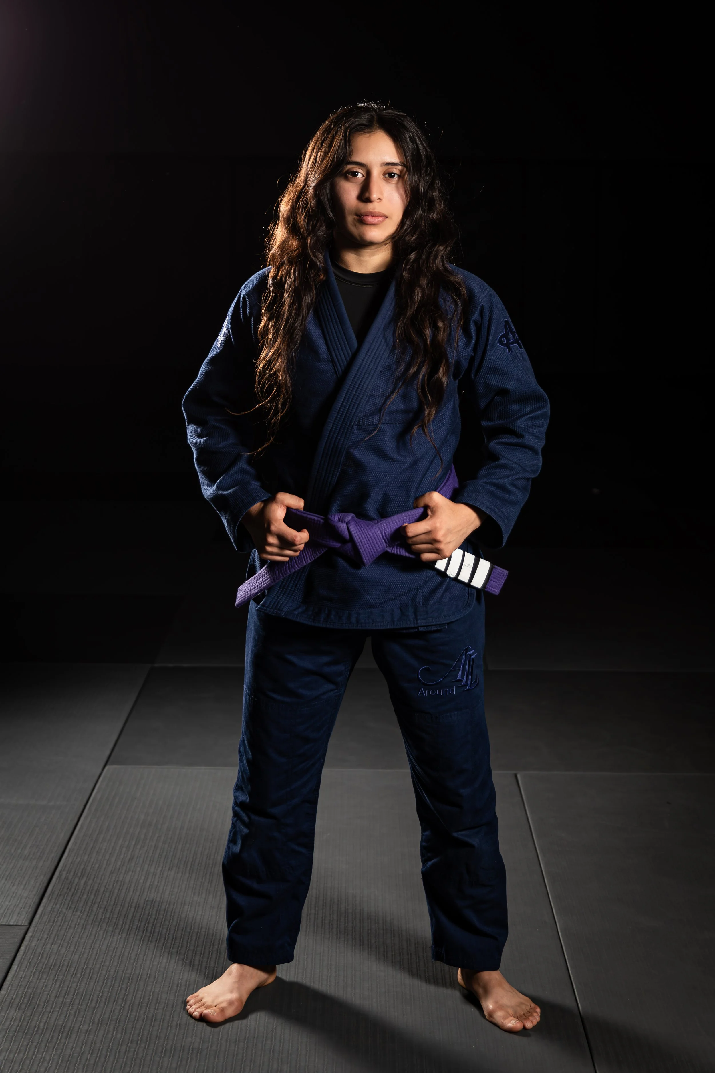 A woman in a blue Brazilian Jiu-Jitsu gi standing on a gray martial arts mat, with her hands on her hips and a purple belt around her waist.