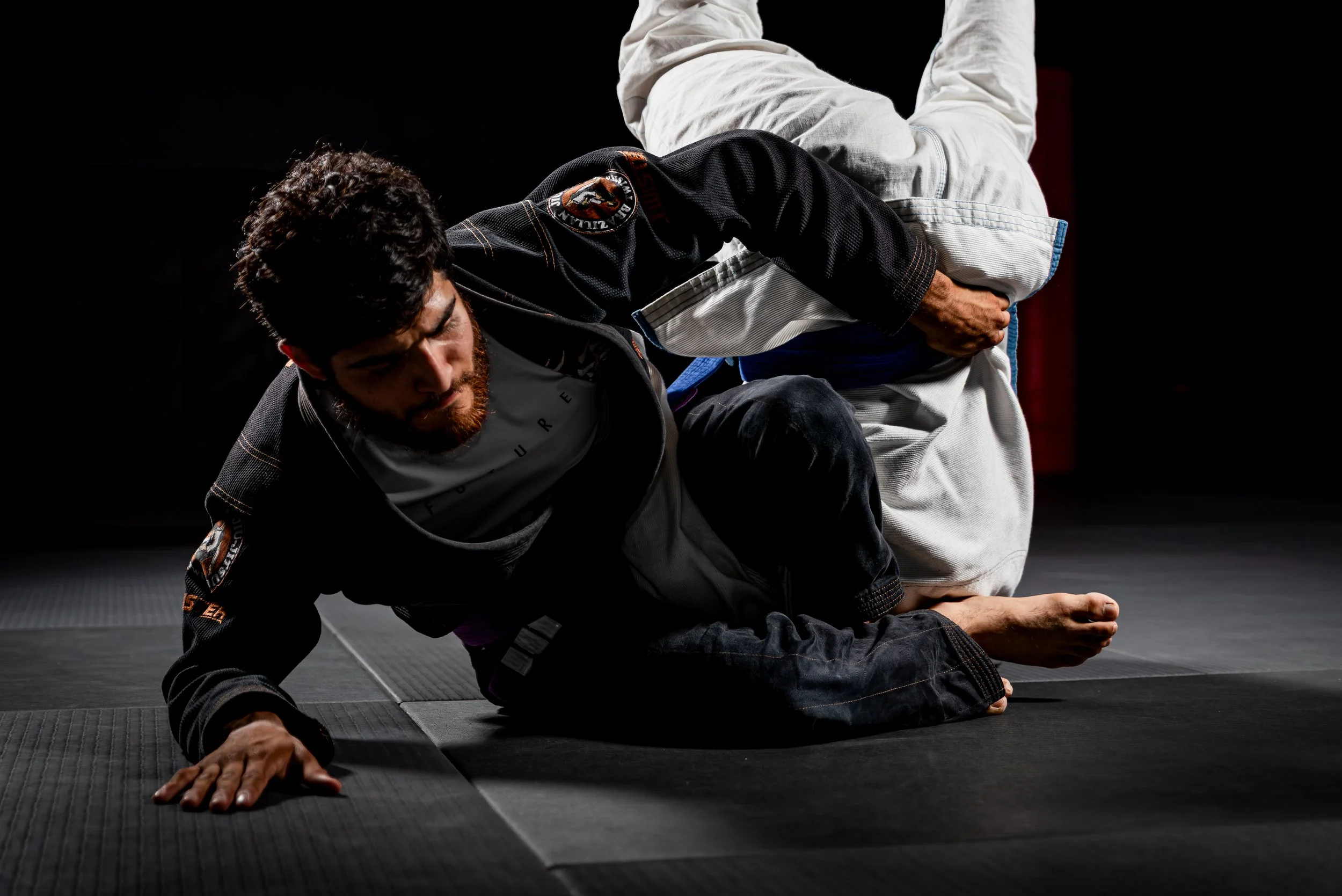 Two martial artists practicing Brazilian Jiu-Jitsu on a black mat; one is executing a technique on the other.