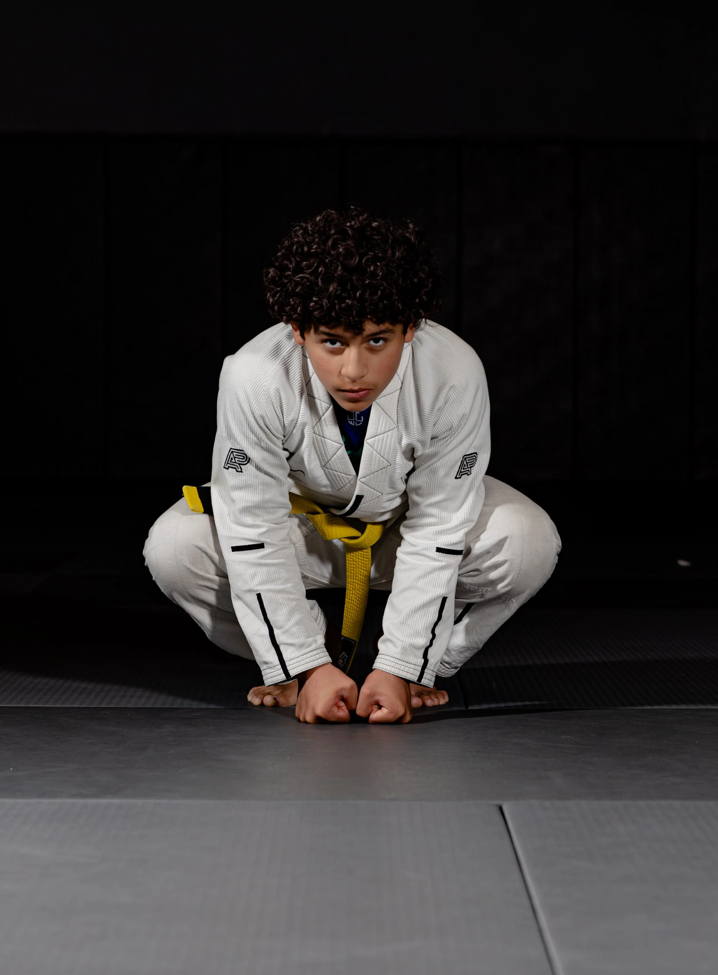 Young person with curly hair in a white gi and yellow belt practicing Brazilian Jiu-Jitsu on a black mat against a black background.