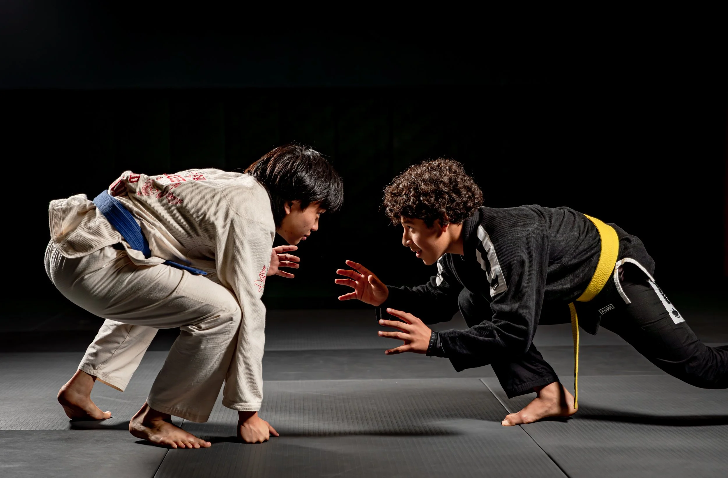 Two martial artists, one in a white gi and the other in a black gi, are practicing Brazilian Jiu-Jitsu on a black mat, facing each other. They are in a low stance with bent knees, hands extended forward, and are focusing intensely on each other.