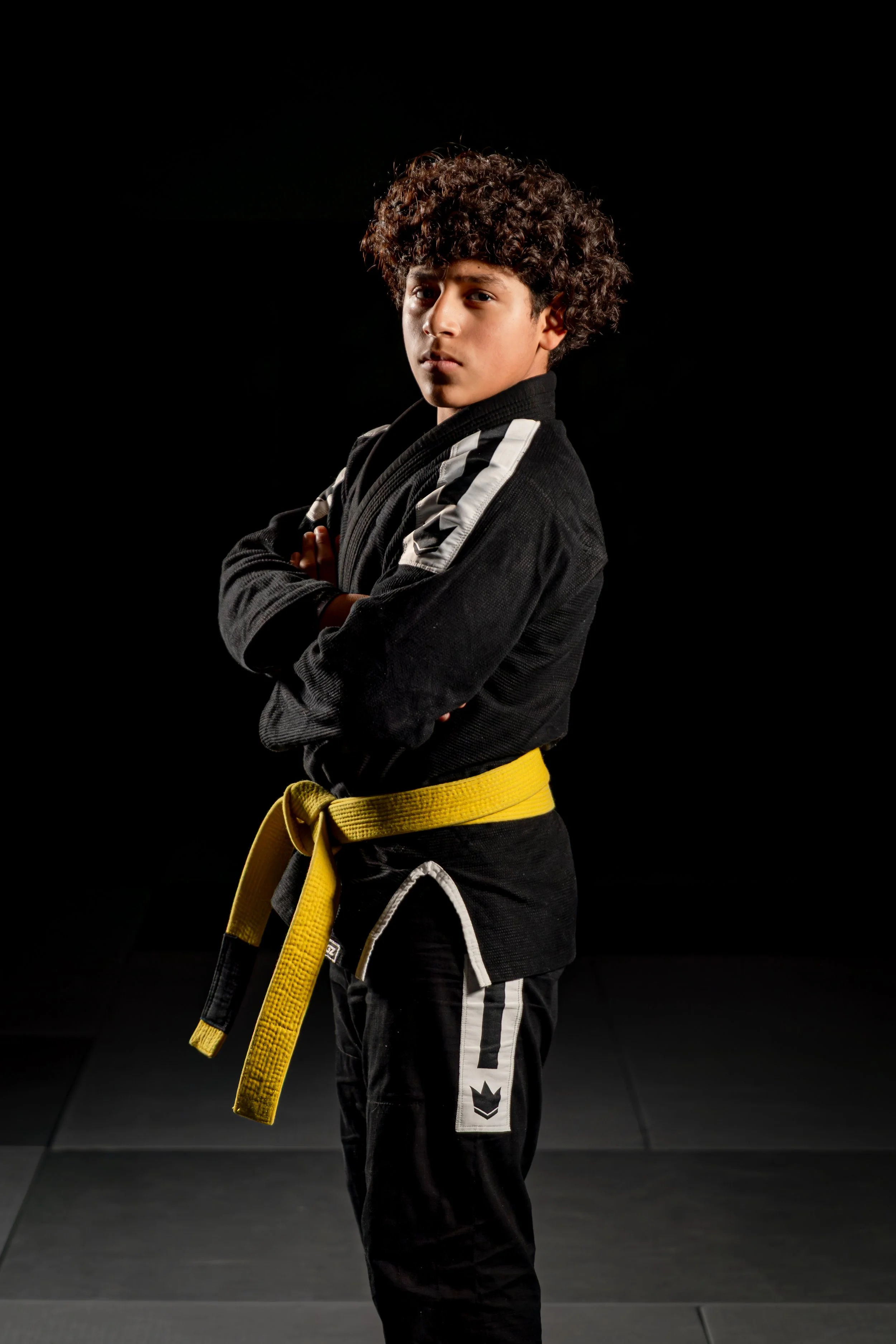 A young person with curly hair standing cross-armed in a black martial arts uniform with yellow belt in a dark training room.