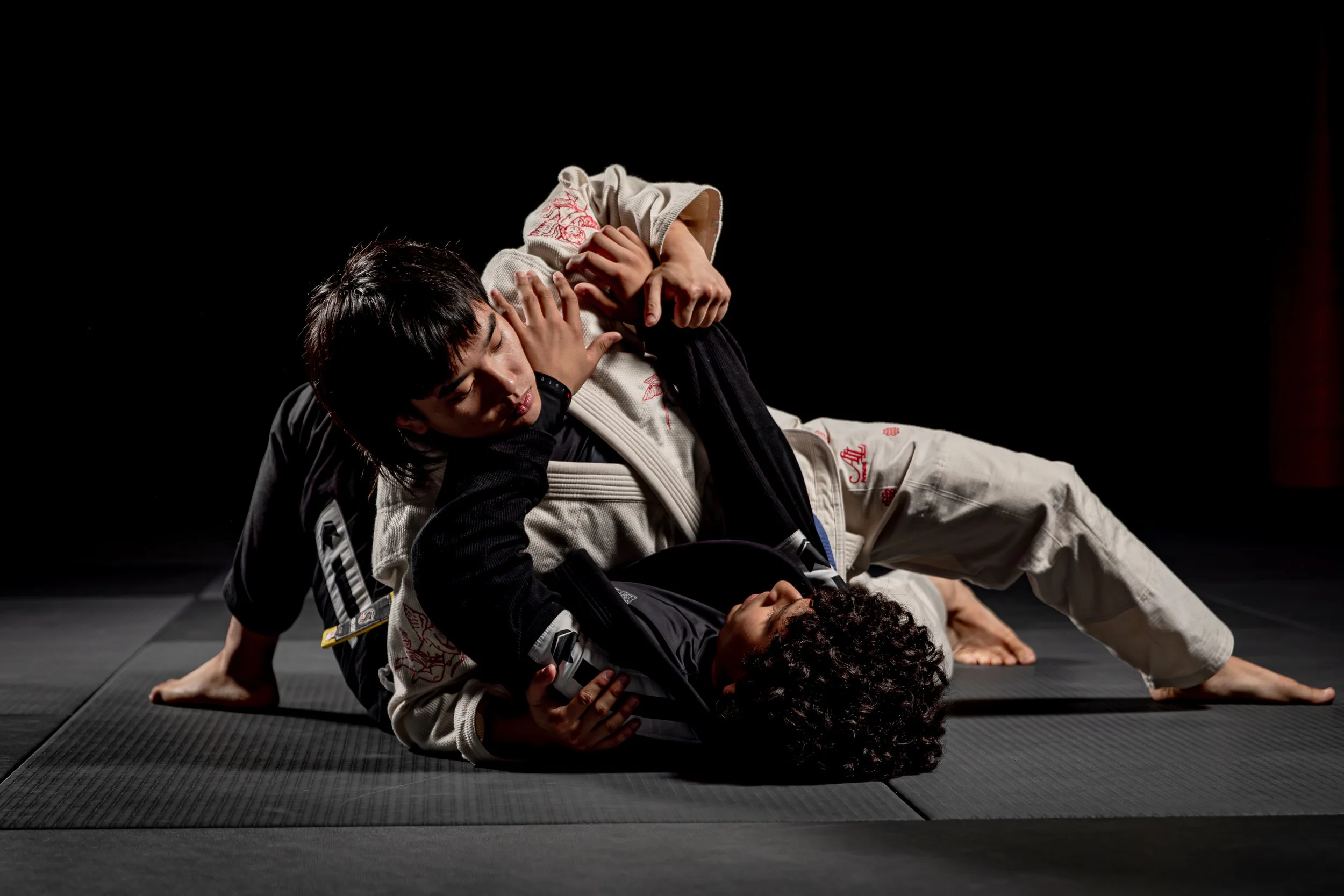 Two Brazilian Jiu-Jitsu practitioners training on a black mat with dark background, one in white gi and the other in black gi, locked in a grappling position, with the person in white on top.