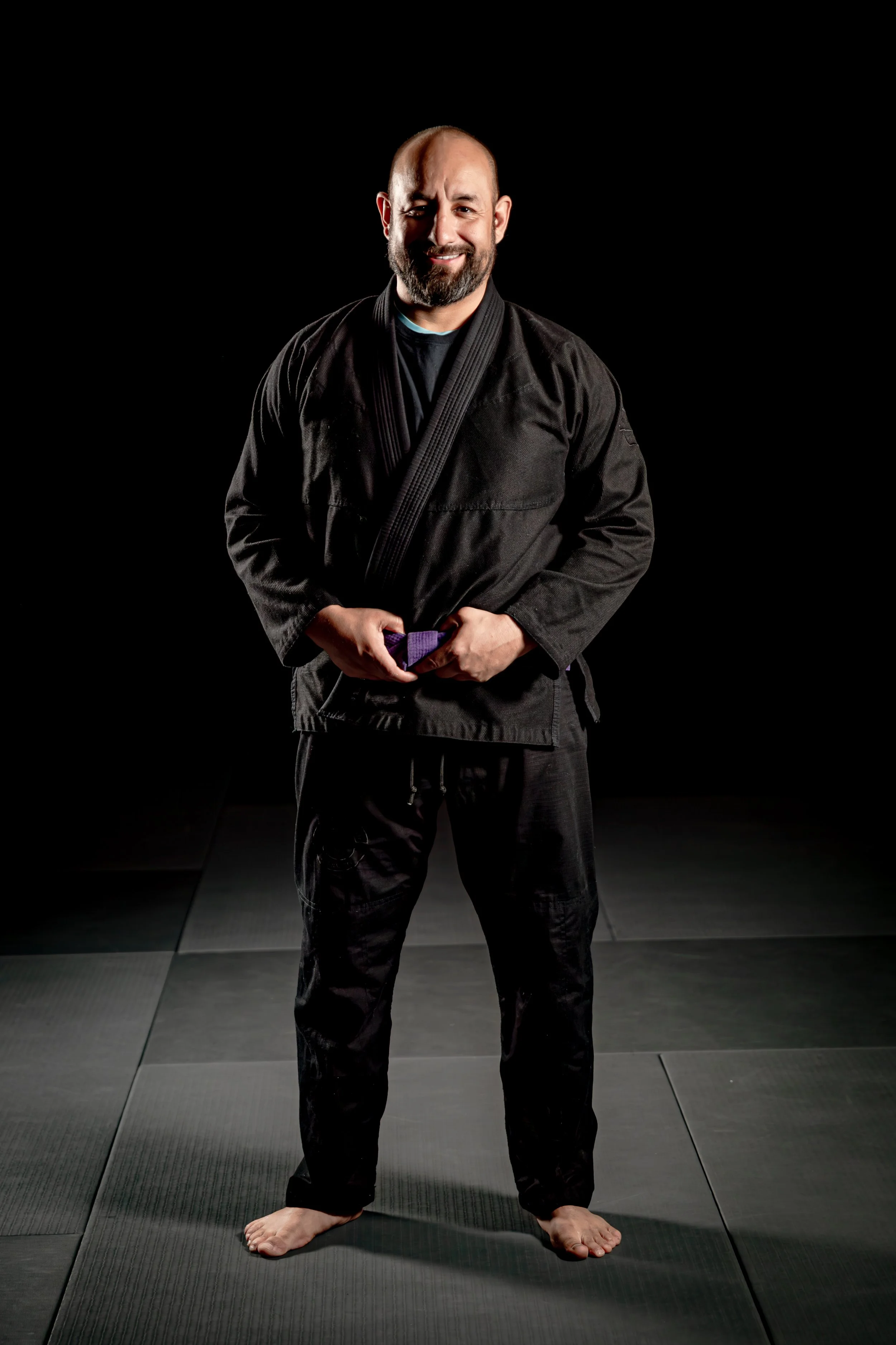 A man in a black martial arts gi standing on a padded mat in a martial arts training area, smiling, holding his fingers together at his waist, with his bare feet on the mats, against a black background.