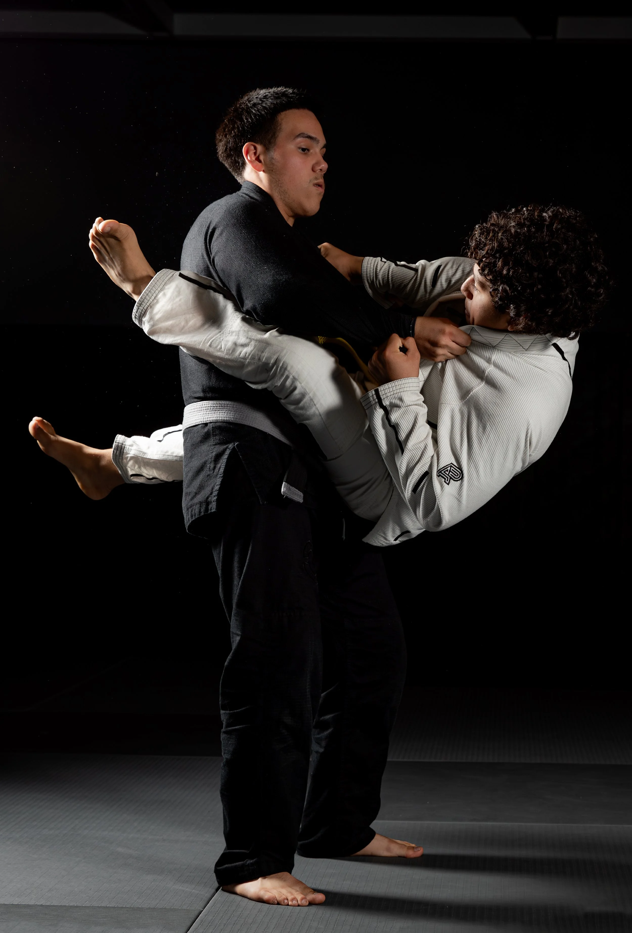 Two martial artists practicing a grappling technique on a black mat, with one person lifting the other. Both are dressed in gis, with one in black and the other in white, in a dark, minimalist training environment.