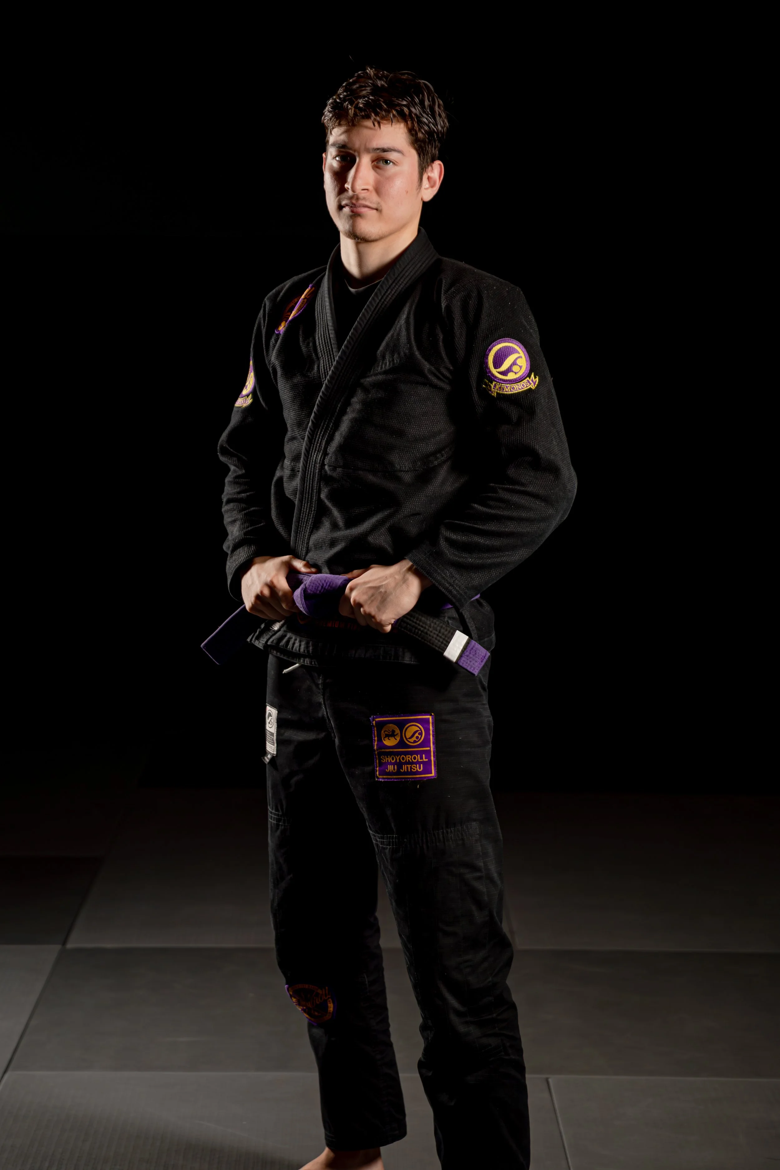 A young man wearing a black Brazilian Jiu Jitsu gi with patches and a purple belt, standing on a martial arts mat in a dark studio.