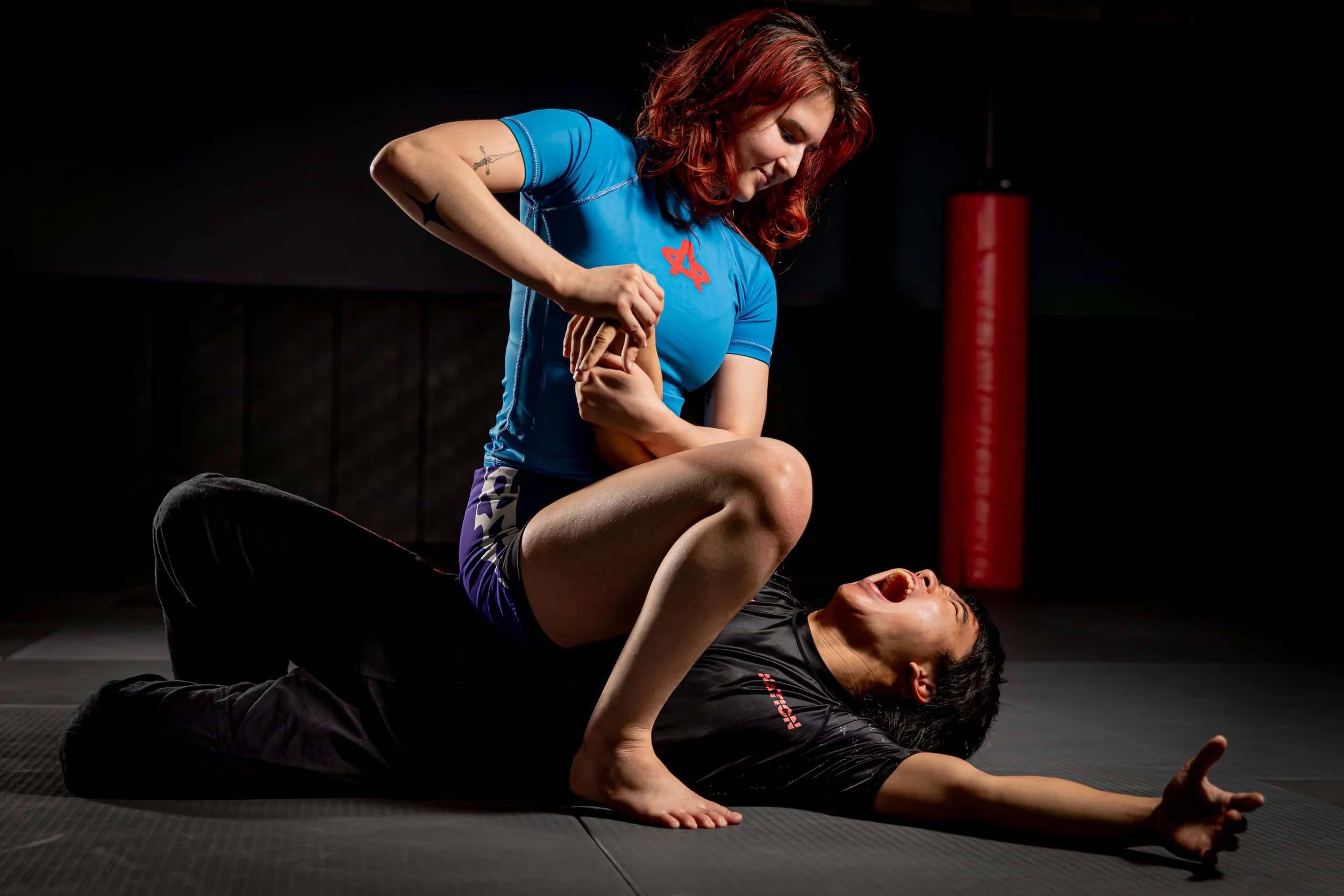 Two women practicing Brazilian Jiu-Jitsu on a black mat, one woman in a blue rash guard on top of the other woman in a black gi during a sparring session in a gym with a red punching bag in the background.