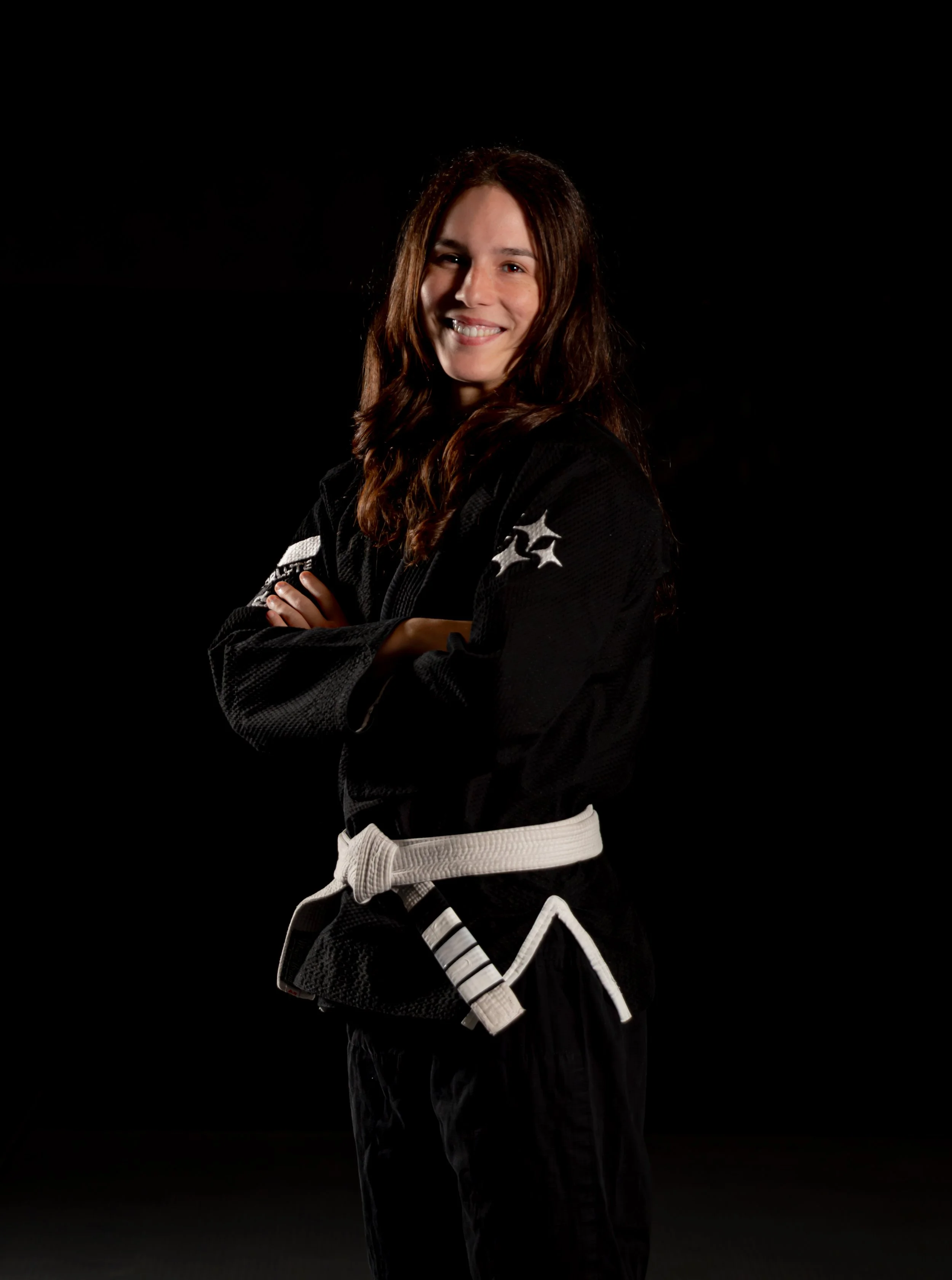 A smiling woman in a black Brazilian Jiu-Jitsu gi with white belt stands with arms crossed against a black background.