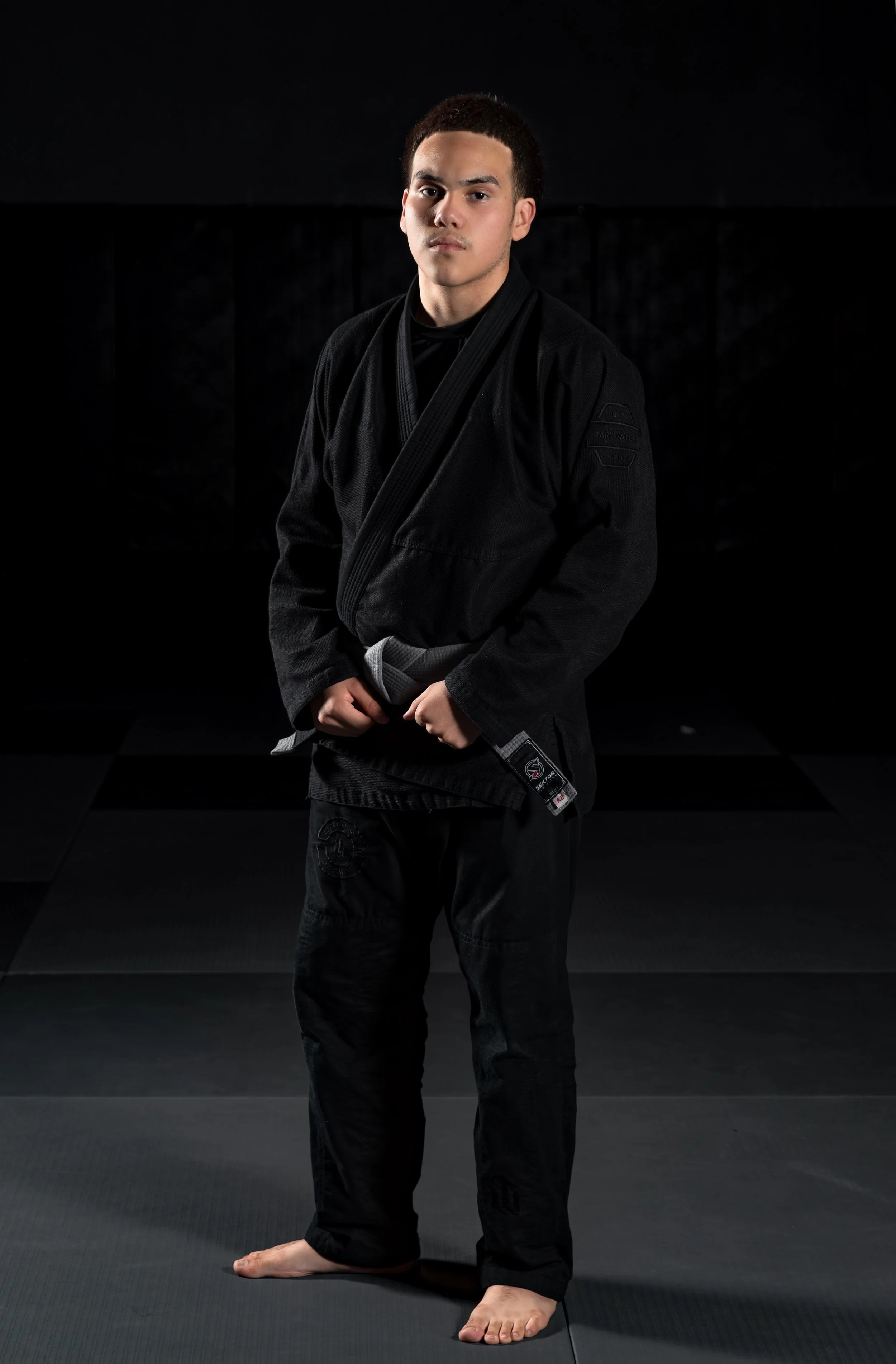 A young man in a black martial arts gi standing on a black mat in a training room with black walls, barefoot, looking directly at the camera.