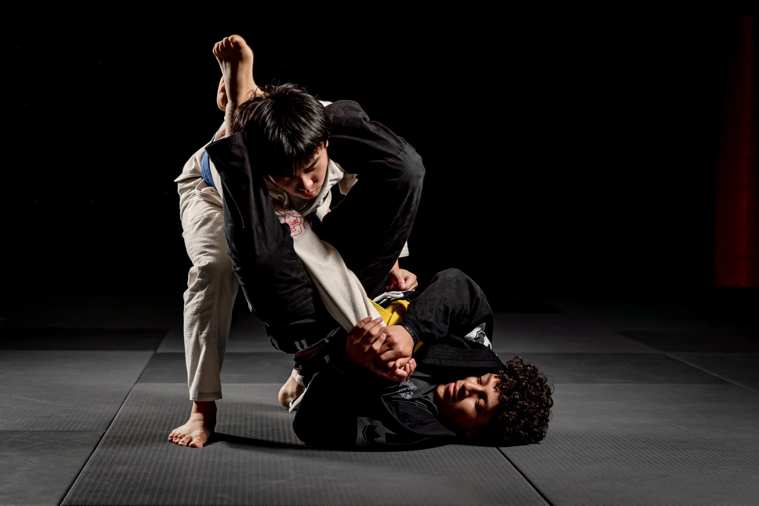 Two martial artists practicing Brazilian Jiu-Jitsu on a black mat in a dimly lit studio. One person is on the ground, smiling, while the other is applying a choke hold from behind.