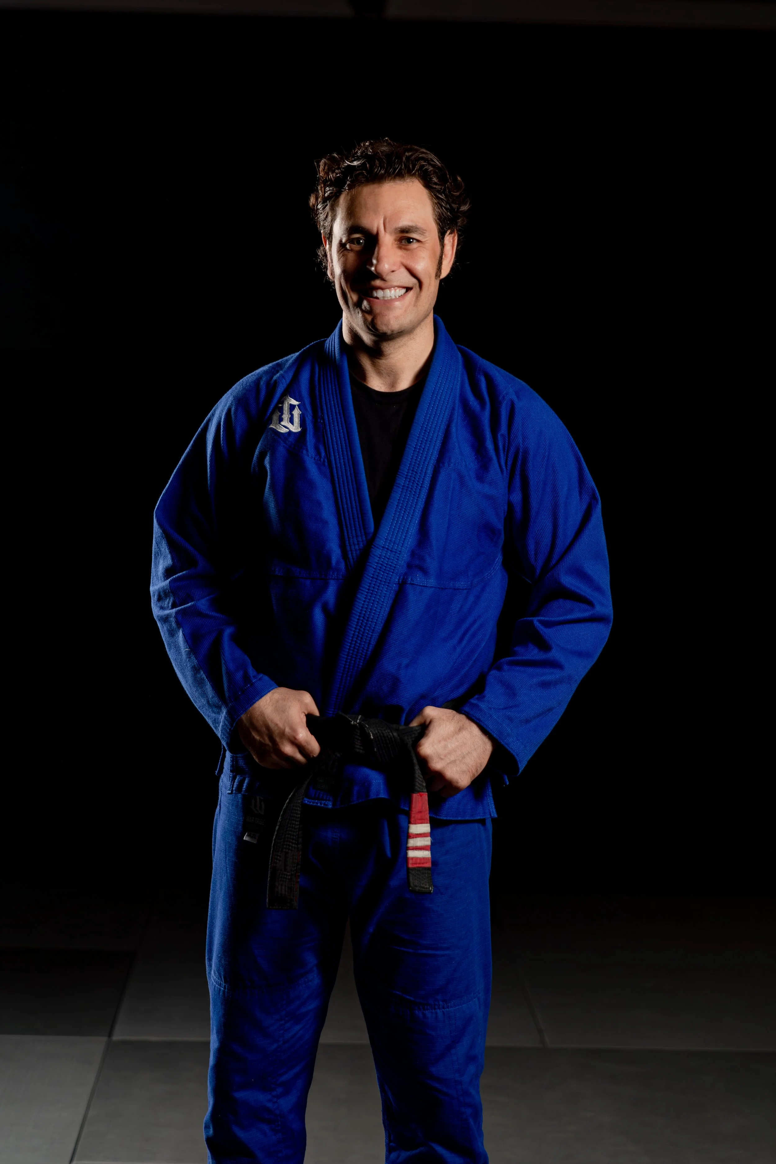 A man in a blue Brazilian jiu-jitsu gi holding a black belt with a red patch, standing on a black mat, smiling at the camera.