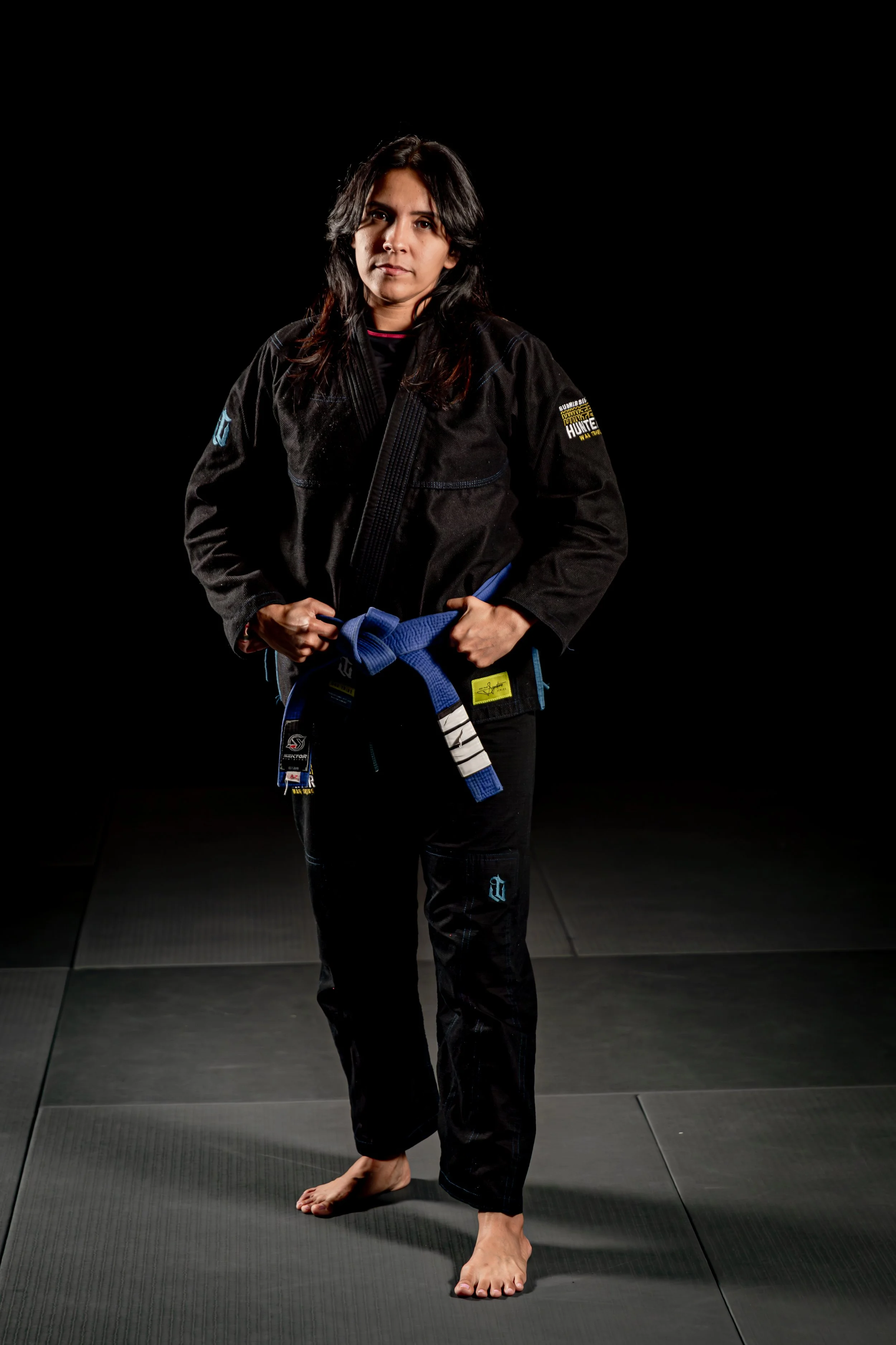 Woman in a black martial arts gi standing on a black mat in a martial arts gym, holding a blue belt, with a serious expression, dark background.