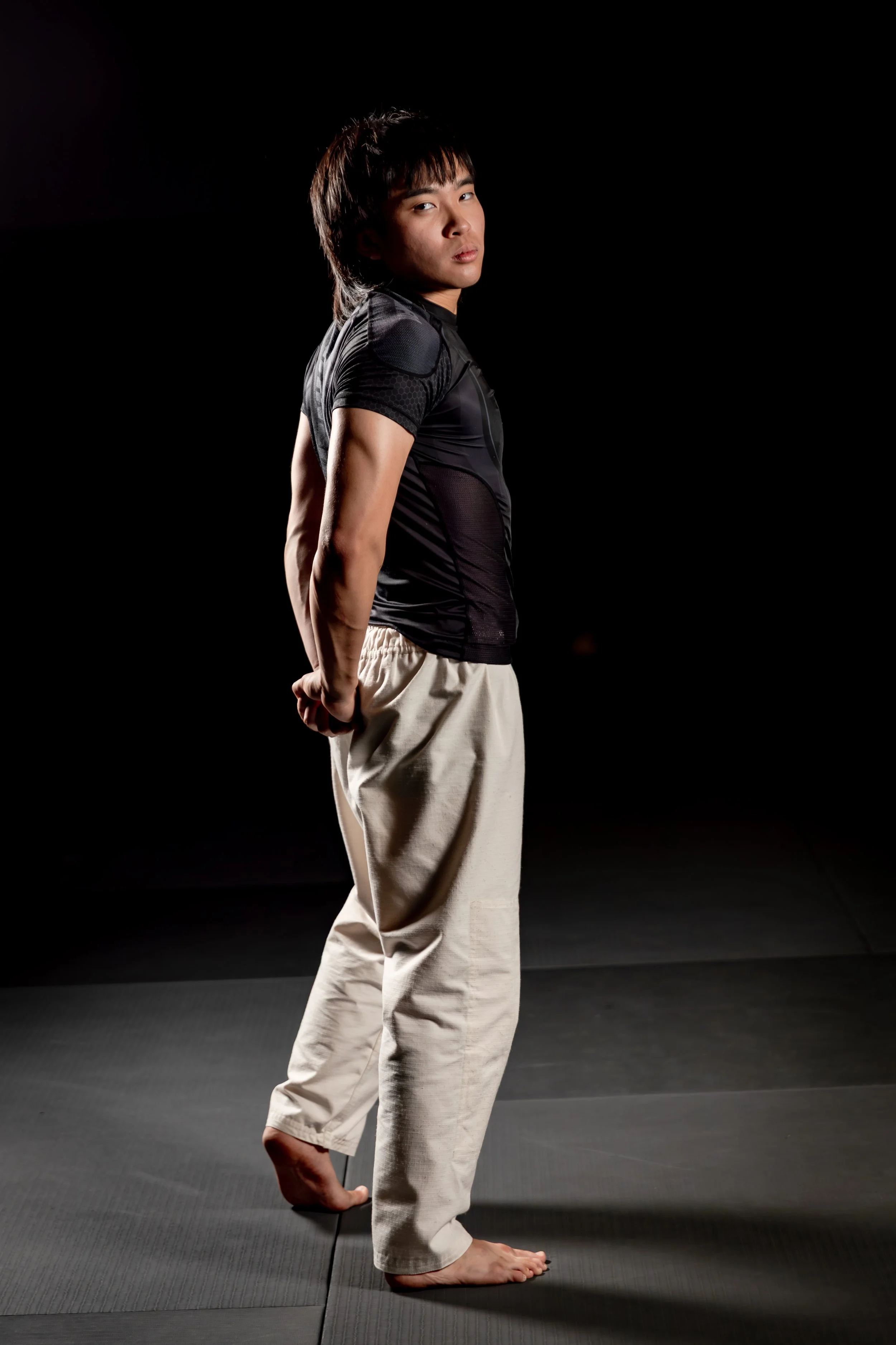 A person standing in a martial arts stance on a black mat, wearing martial arts uniform pants and a black athletic shirt, with a dark background.