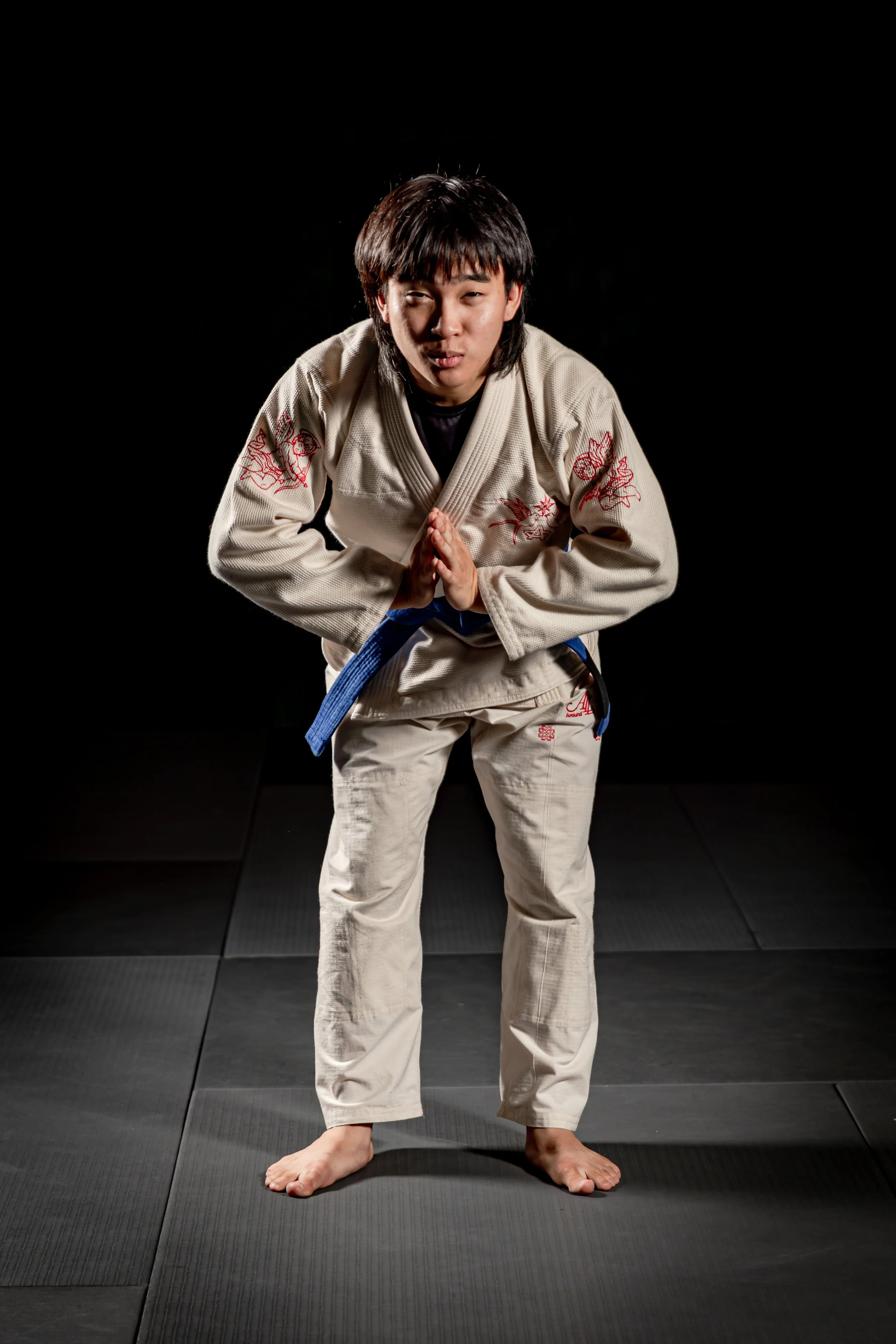 Young woman in a martial arts gi with a blue belt, standing on a black mat, in a bowing pose with hands pressed together.