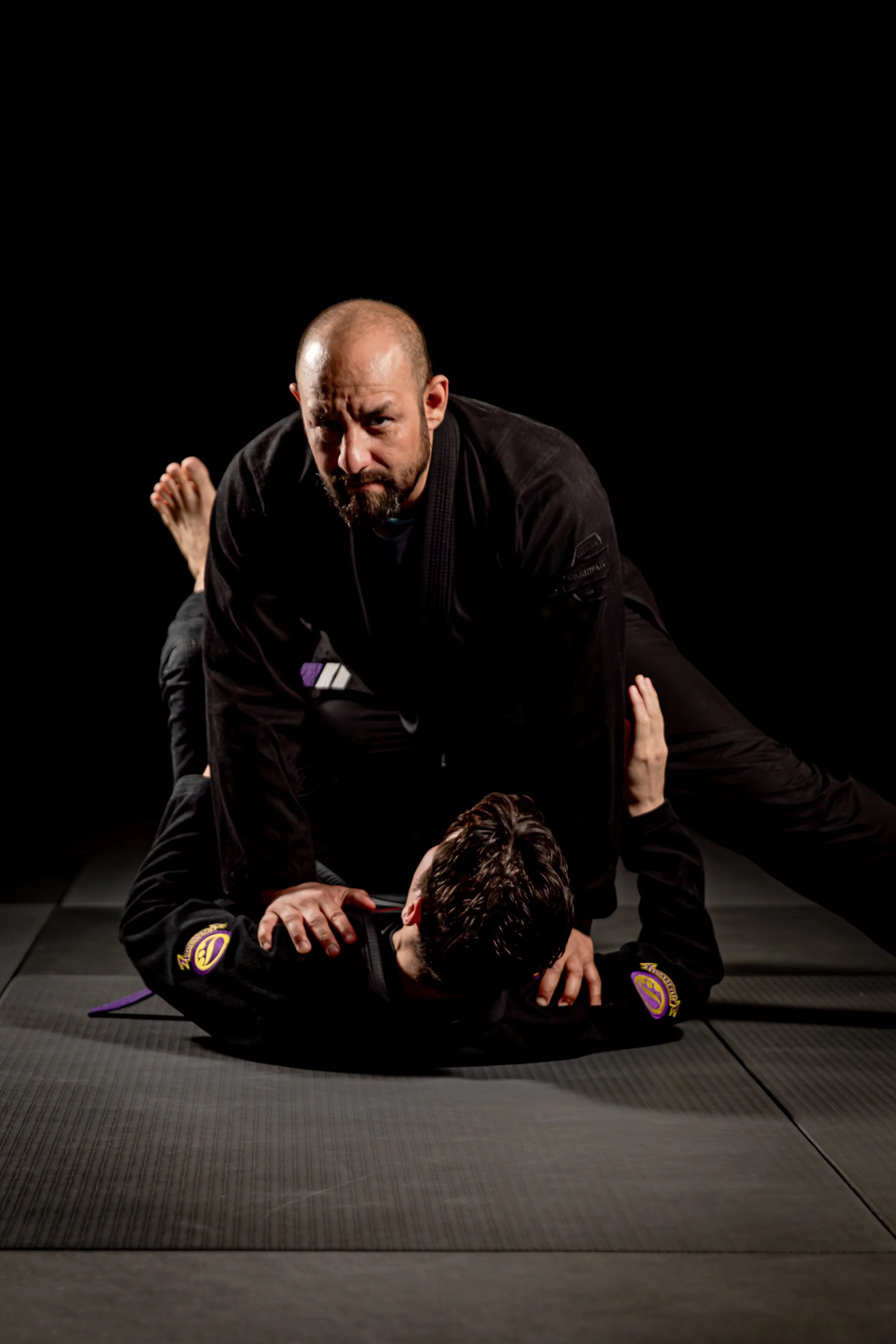 Martial arts training with an instructor demonstrating techniques on a student in a black gi, on a black mat with a dark background.