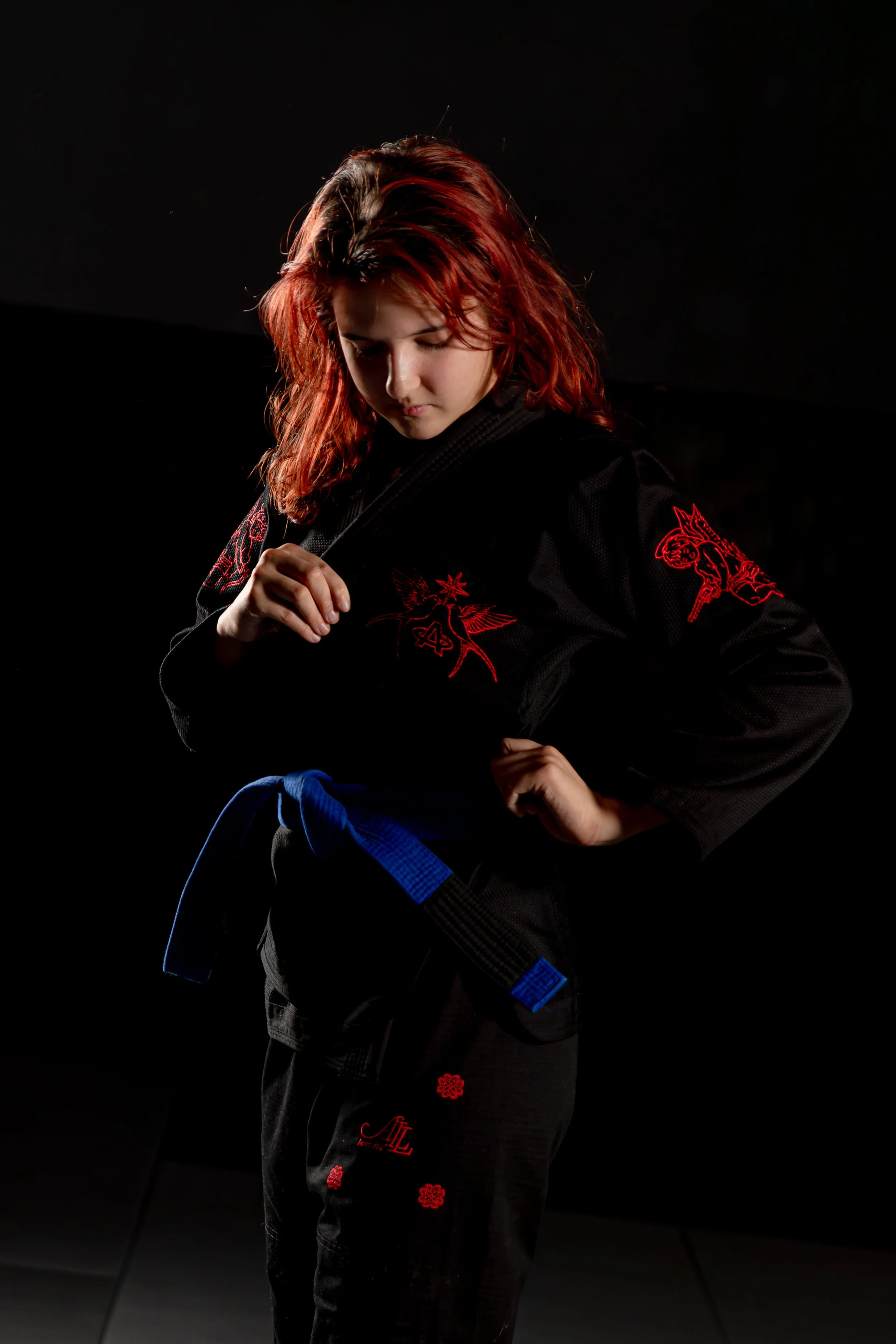 A young woman with red hair wearing a black martial arts gi with red embroidery, holding a blue belt, in a dark setting.