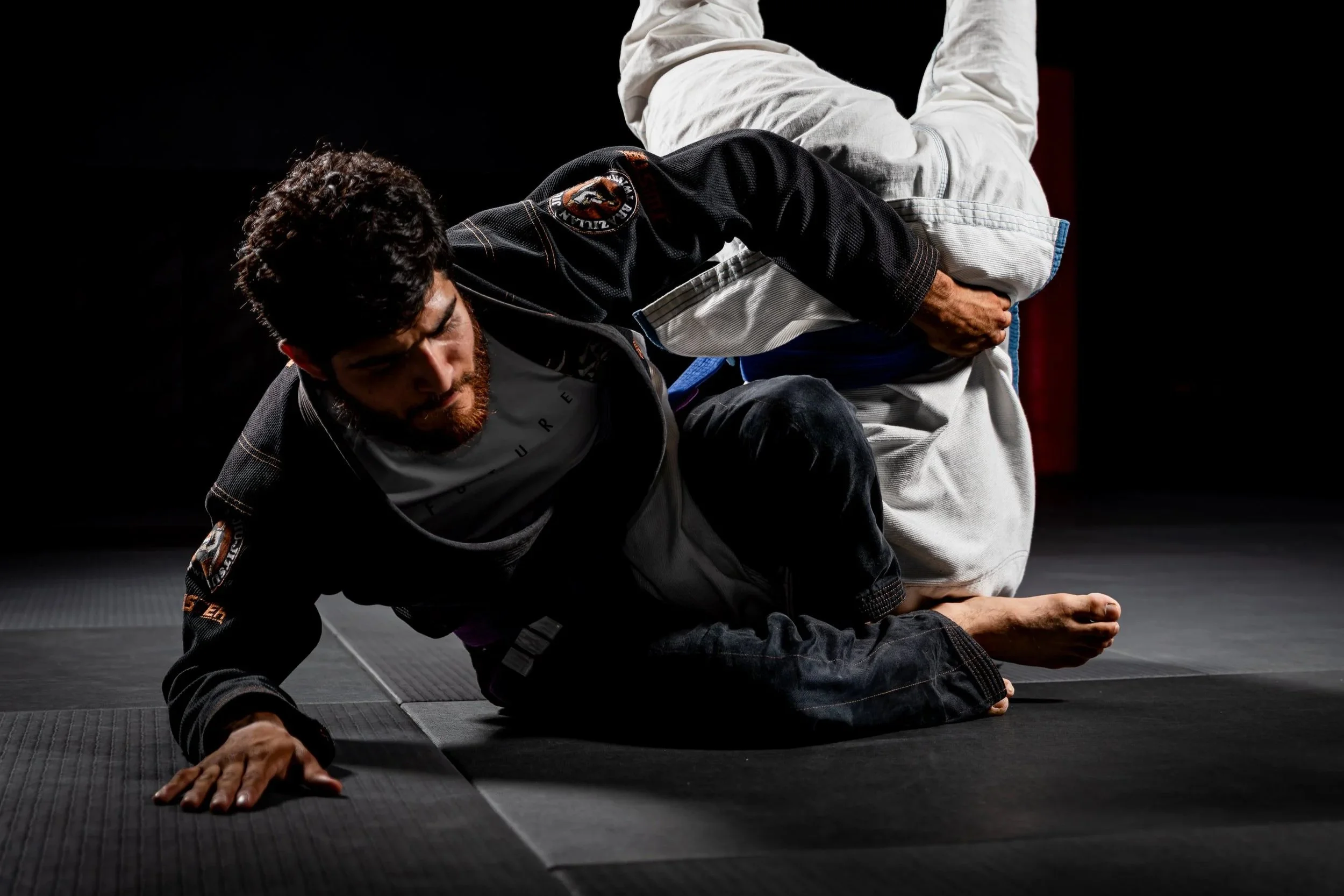 Two martial artists demonstrating Brazilian Jiu-Jitsu techniques on black mats in a dark training area.