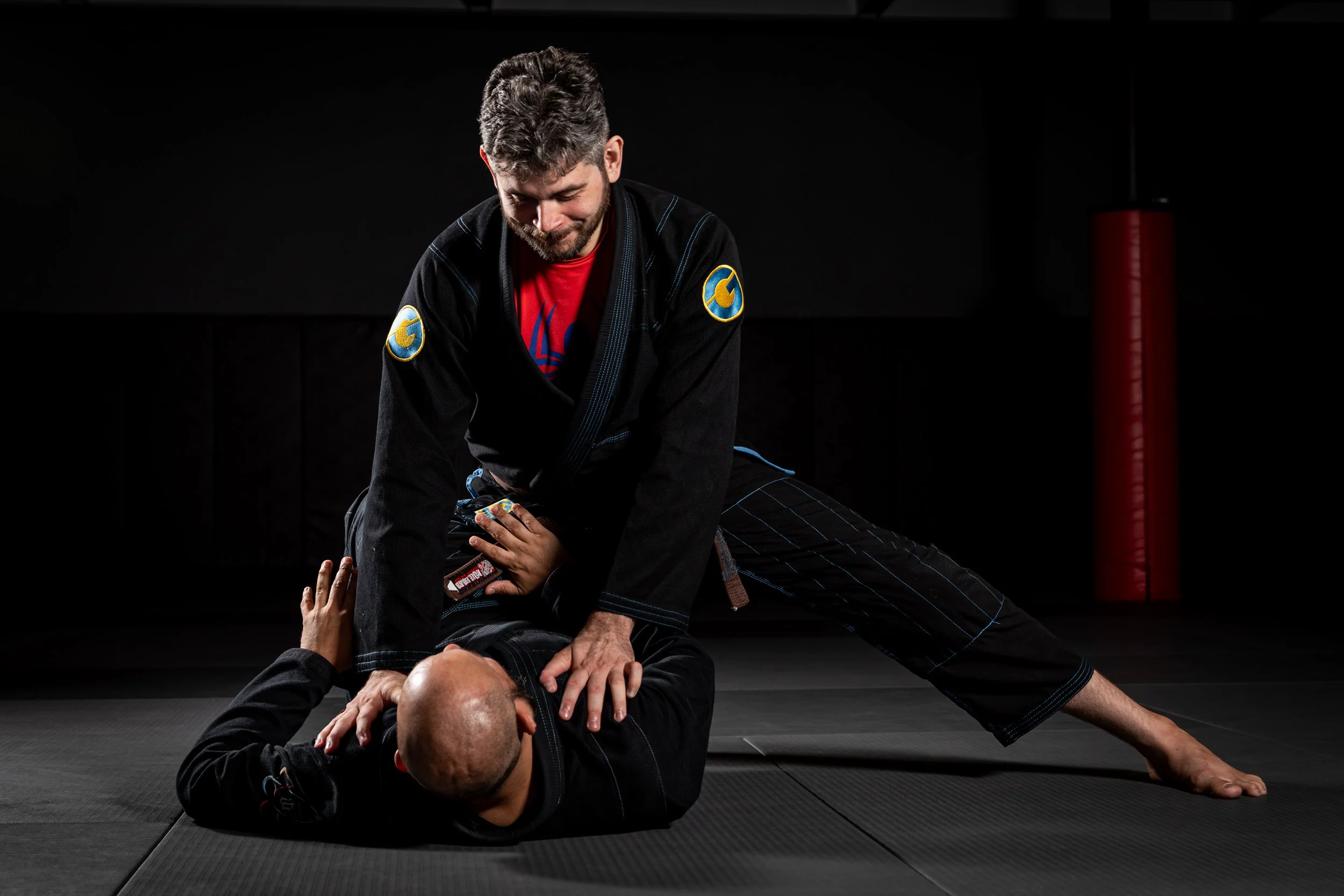 Two men practicing Brazilian Jiu-Jitsu on a black mat, with one man in a black gi on top of another man in a black gi, in a dark training room.