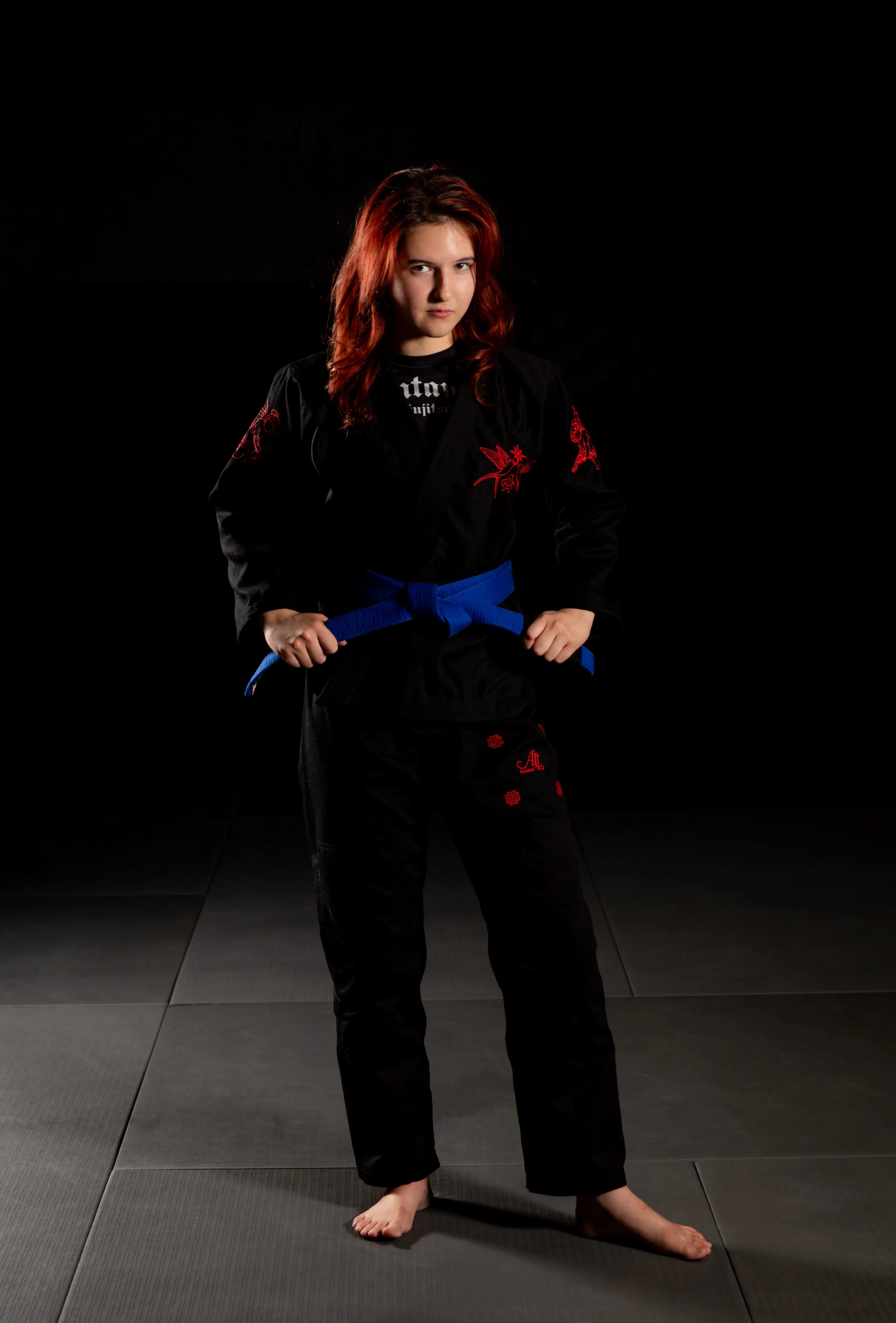 Woman wearing a black Brazilian Jiu-Jitsu gi with red and blue embroidery, standing on a dark martial arts mat, holding a blue belt, with one foot forward and a serious expression.