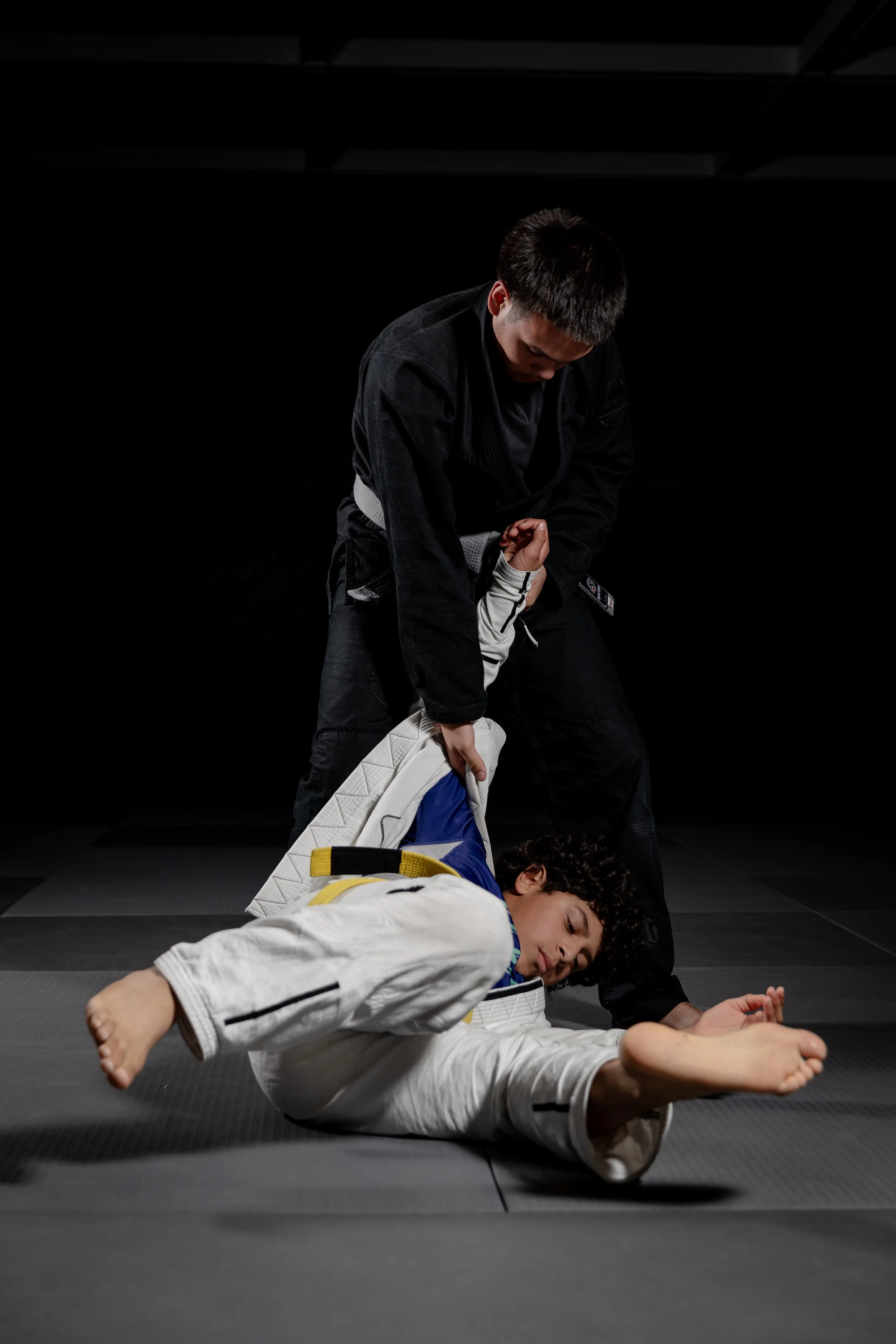 Two martial artists practicing Brazilian Jiu-Jitsu on a black mat. The person in the foreground, wearing a white gi, is lying on the mat. The person in the background, wearing a black gi, is standing over the person in the foreground, gripping their 