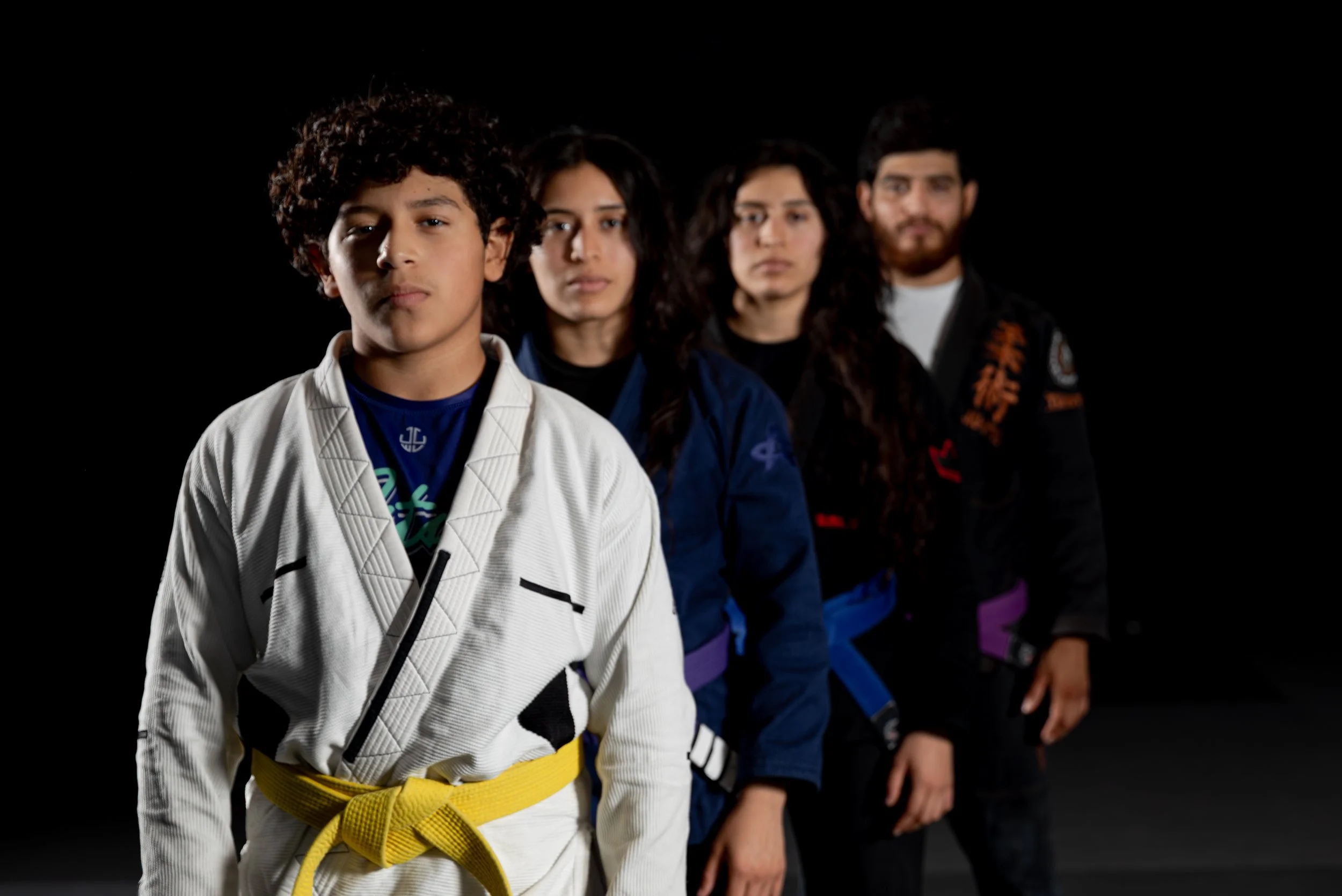 A line of four martial artists wearing martial arts uniforms and colored belts, standing in a black background.