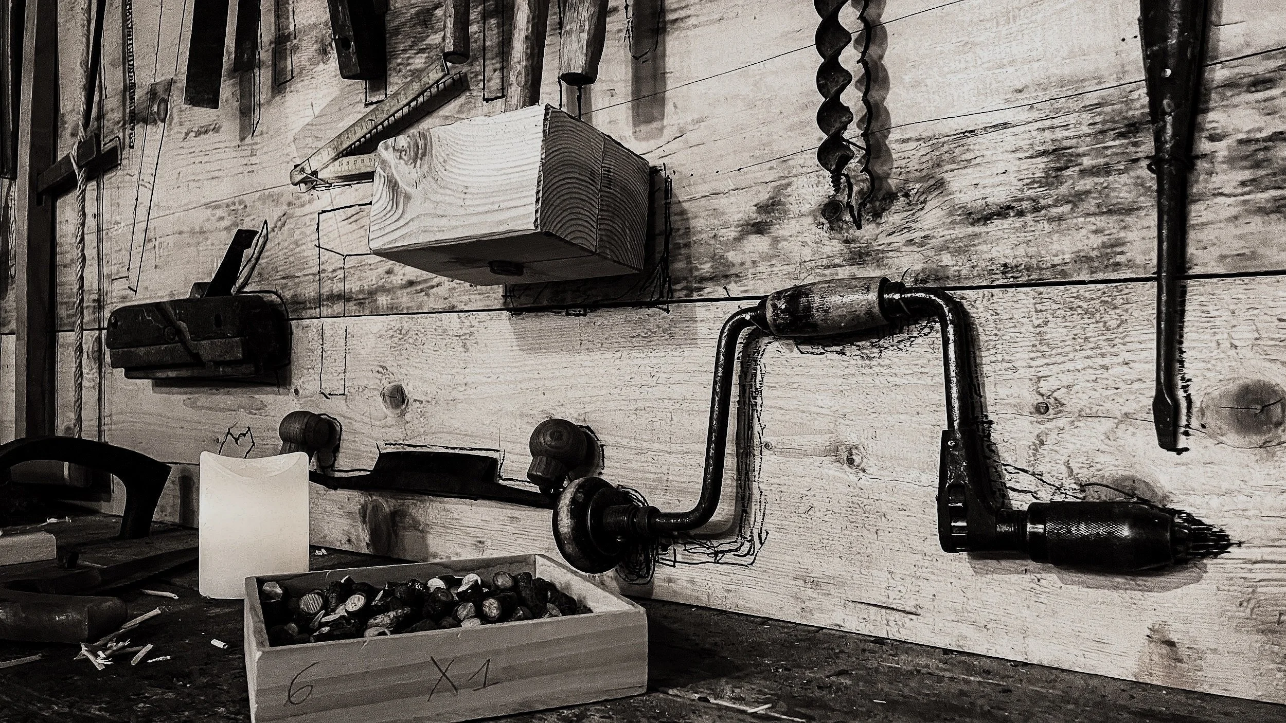 Black and white photo of a rustic workshop wall with tools hanging, including a brace, a block of wood, and a box filled with nails or screws on a work surface.