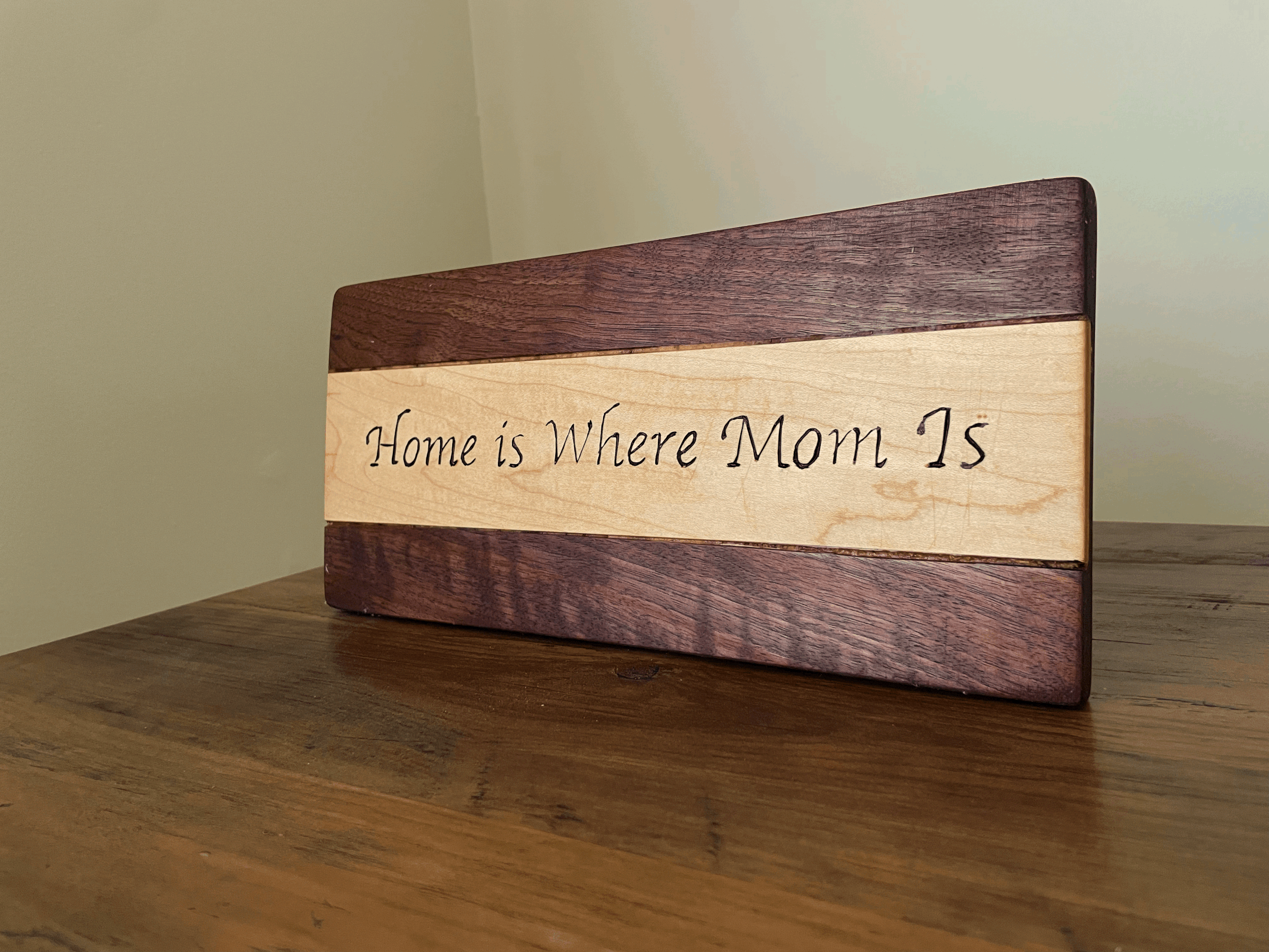 A wooden block with a quote, partially visible, that says, "Home is Where Mom Is," placed on a wooden surface against a plain wall.