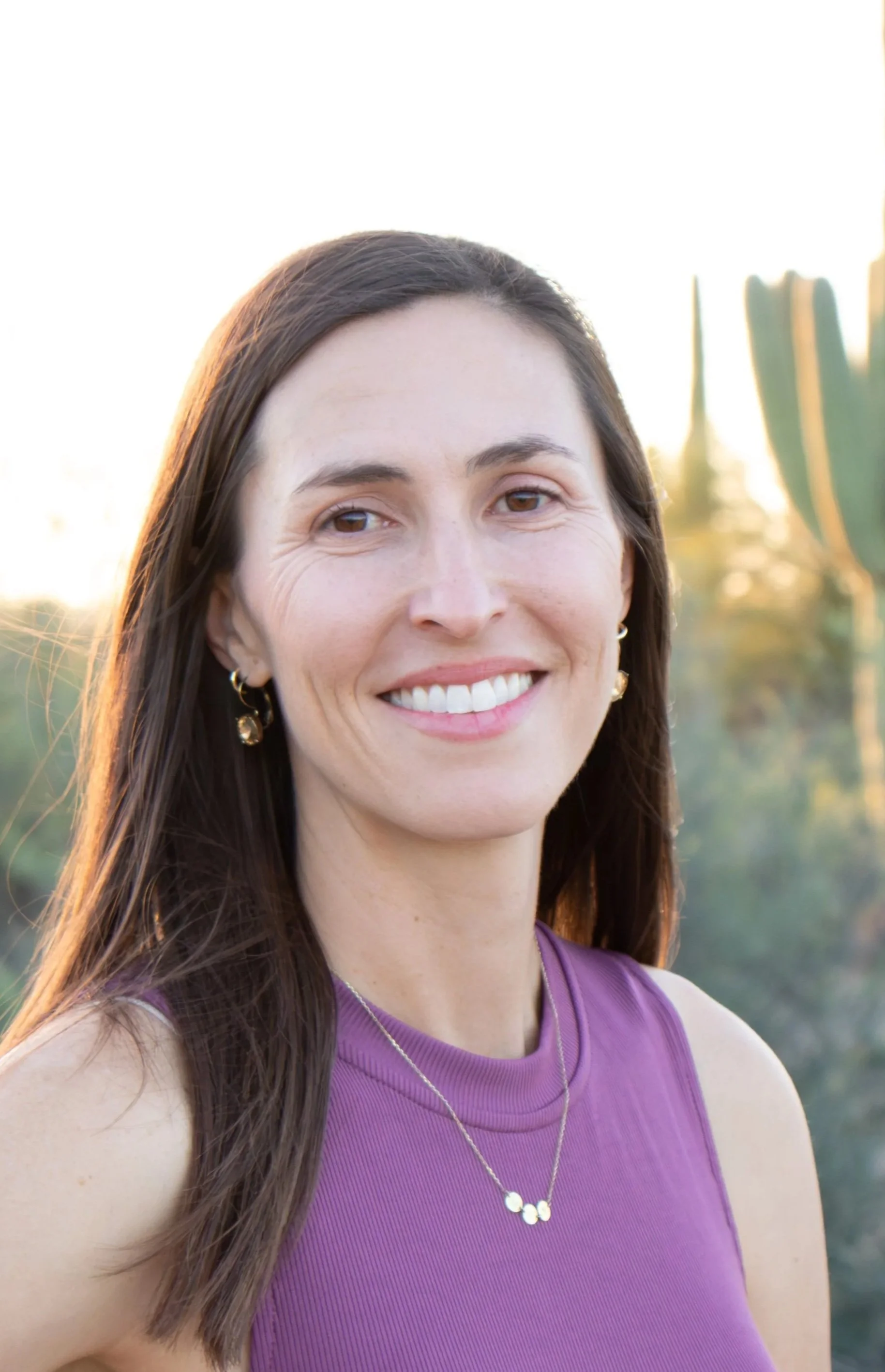 A woman with long brown hair, smiling, wearing a purple sleeveless top, gold earrings, and a gold necklace, standing outdoors with sunlight and cactus plants in the background.