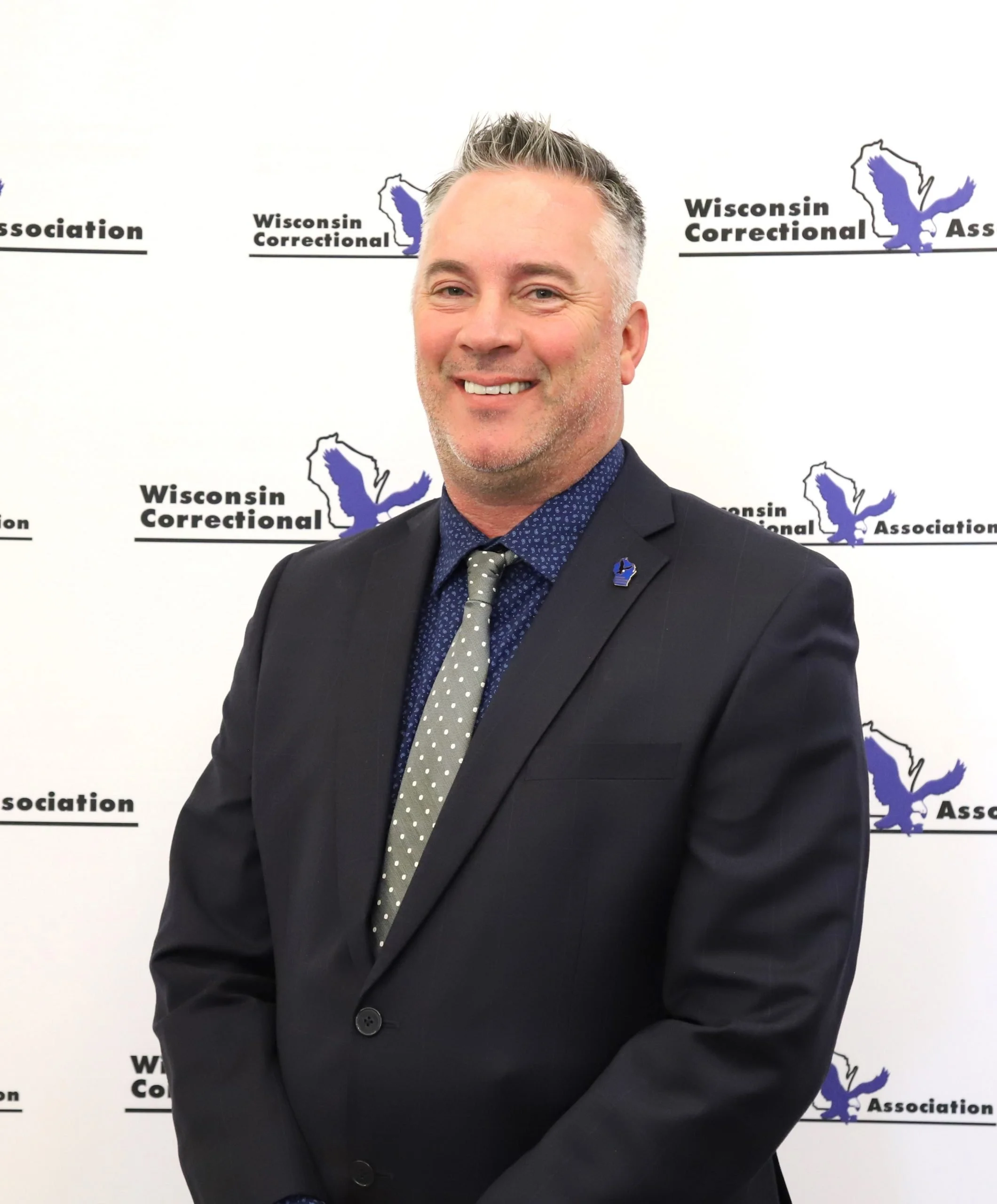 A man in a dark suit with a patterned tie and a pins on his lapel, smiling, standing in front of a backdrop with the Wisconsin Correctional Association logo.