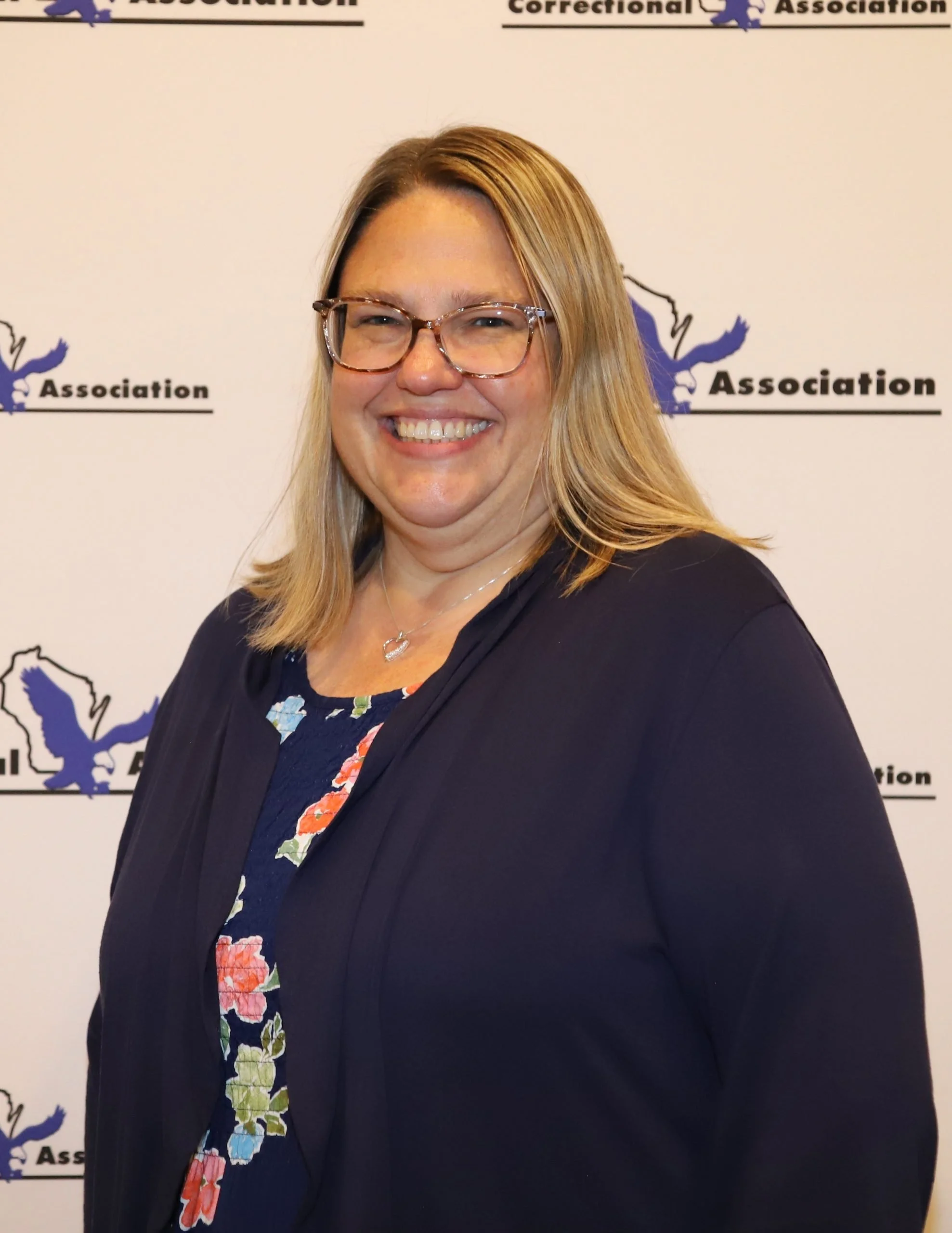 A smiling woman with blonde hair and glasses standing in front of a white backdrop with logos and text related to a correctional association.