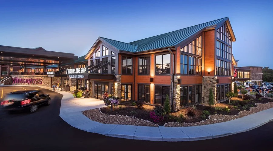 Exterior view of a modern, multi-story building with large glass windows and a green metal roof, surrounded by landscaping and parking lot during the evening.