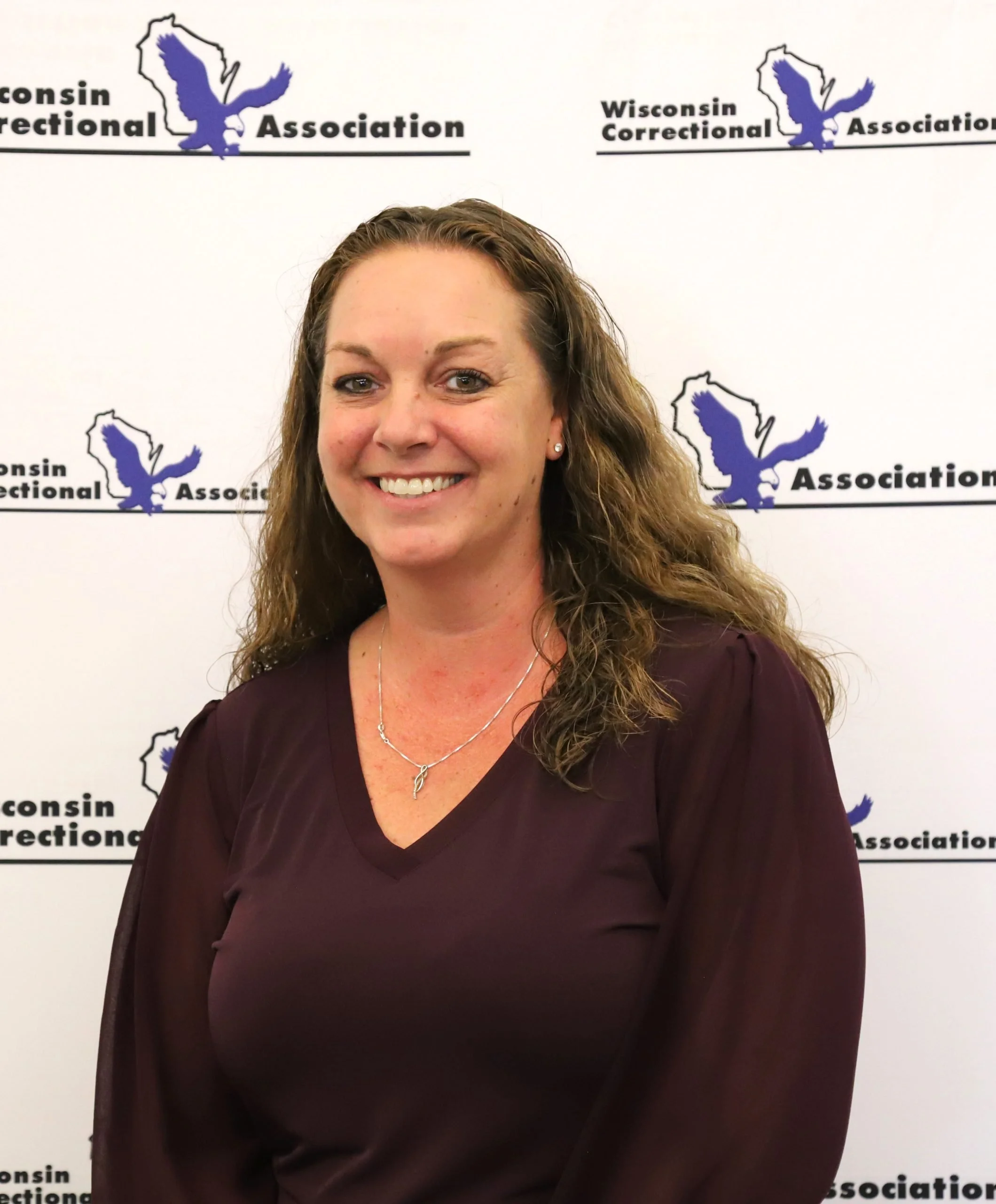 Smiling woman with long curly brown hair standing in front of a background with the Wisconsin Correctional Association logo, wearing a dark purple blouse and a silver necklace.
