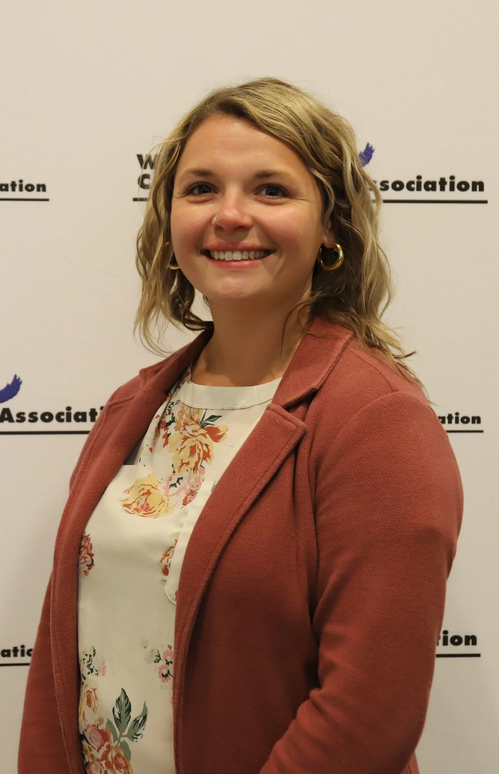 Portrait of a woman with shoulder-length wavy blond hair, wearing hoop earrings, a nose ring, a floral blouse, and a reddish-brown blazer, standing in front of a white backdrop with the logos of the Women's Cable Association.