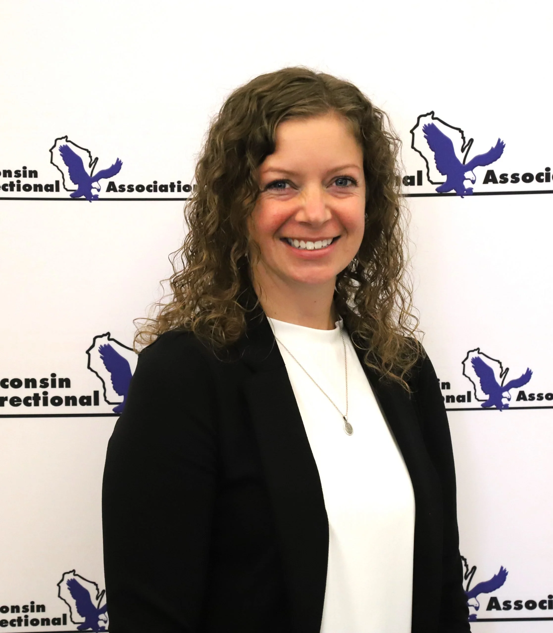 A woman with curly brown hair smiling, standing in front of a white background with the Wisconsin Regional Association logo featuring a blue eagle and the text.
