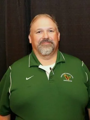 A man with a beard and mustache wearing a green sports polo shirt with a logo, standing indoors against a dark background.