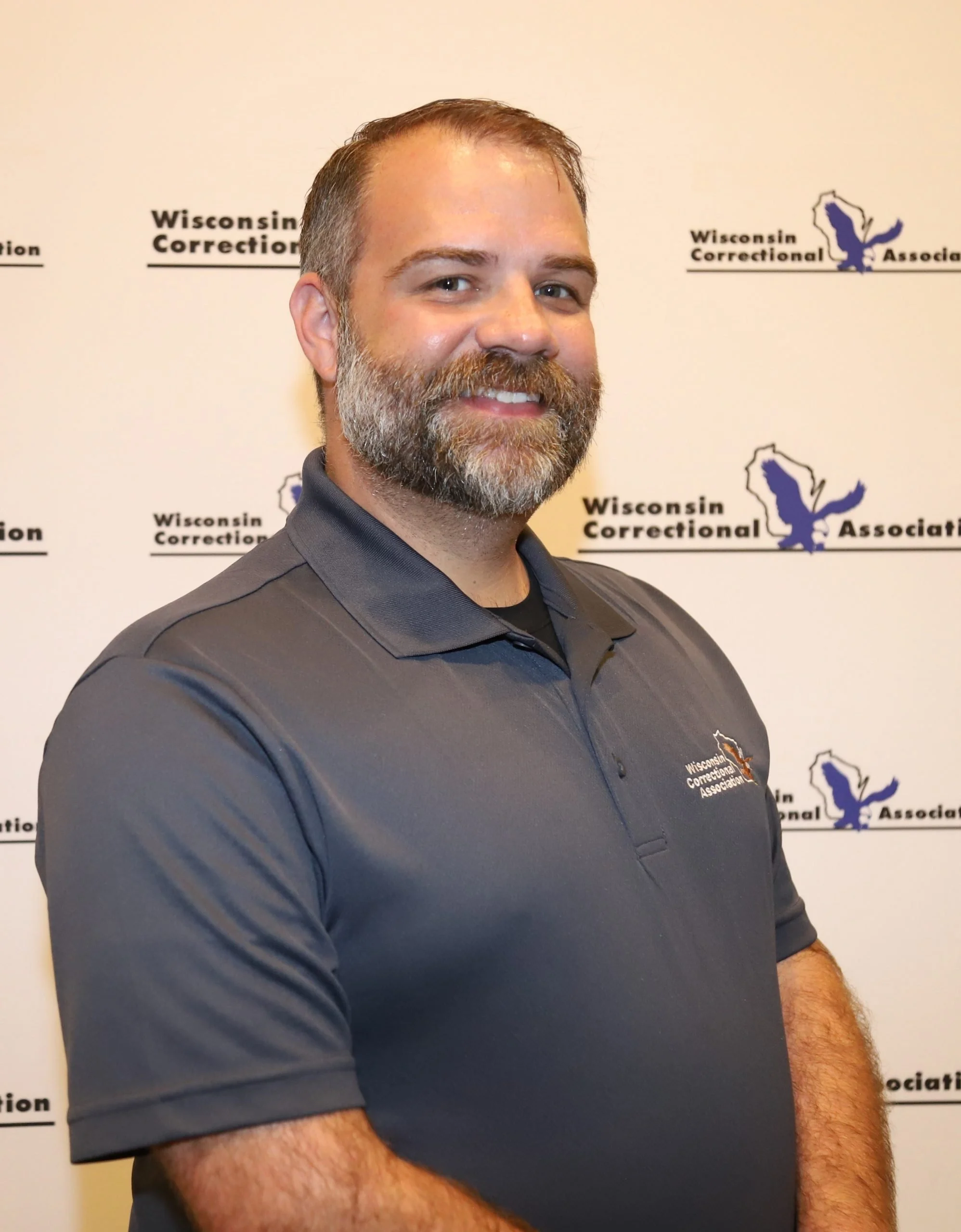 A man with a beard and mustache, smiling, standing in front of a backdrop with the Wisconsin Correctional Association's logo and text.