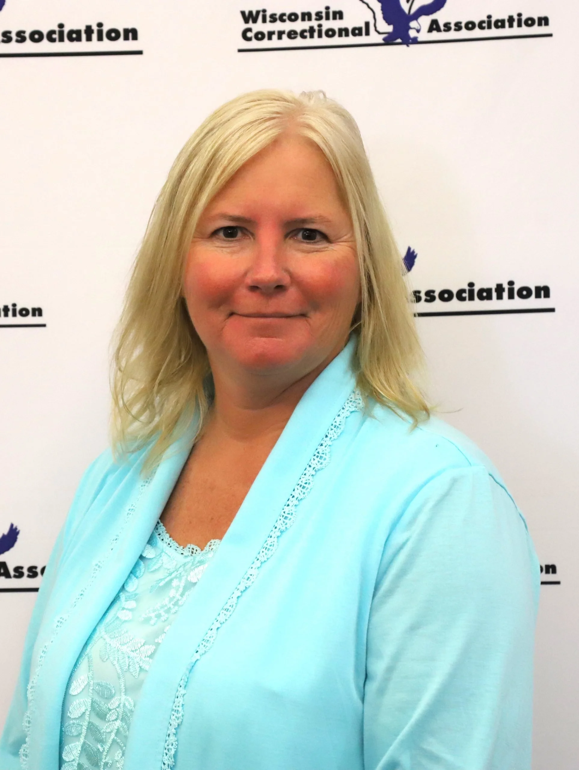 A blonde woman in a light blue jacket and white lace top standing in front of a white backdrop with Wisconsin Correctional Association logos.