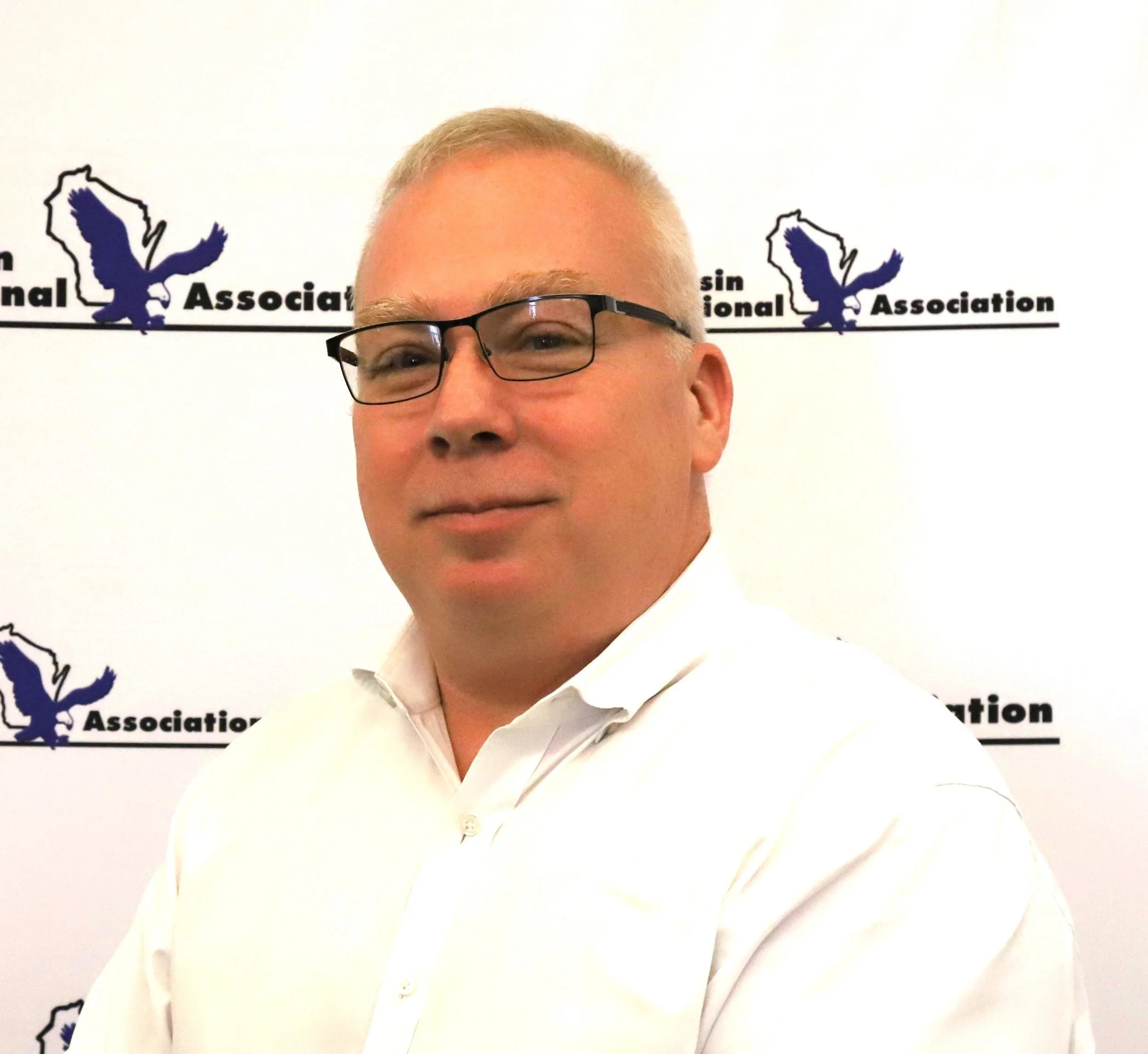 A man with blond hair, glasses, and a white shirt standing in front of a backdrop with the logo and name of the Wisconsin Association.