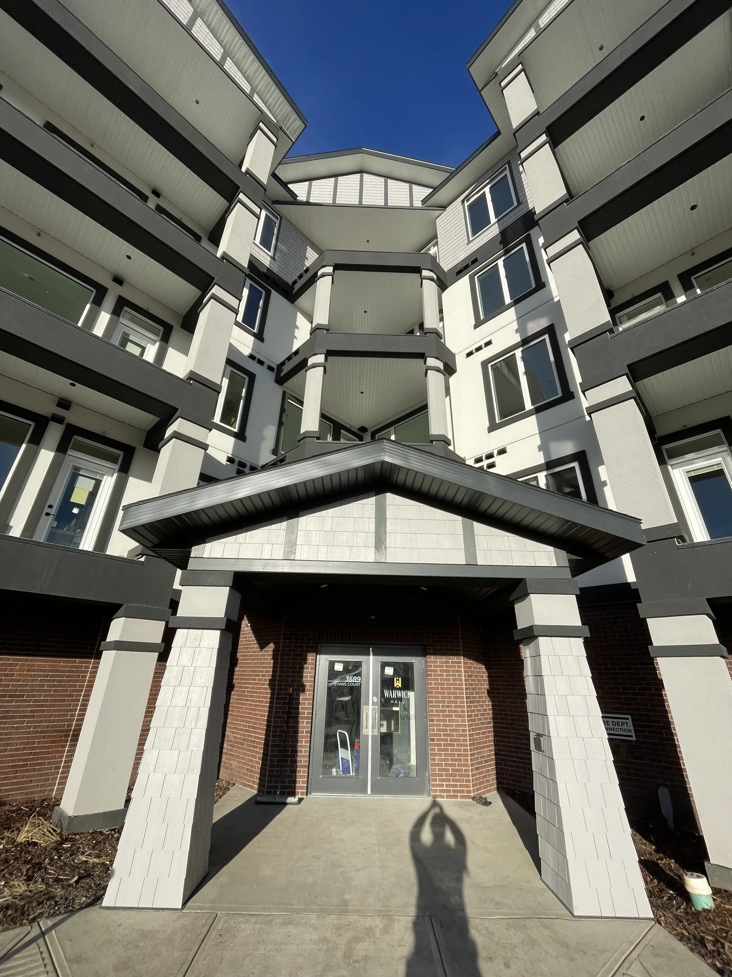 Low-angle view of a modern multi-story apartment building with balconies and glass-paned doors, under a clear blue sky. The entrance has a small overhang, and a person's shadow is visible on the ground in front of the door.
