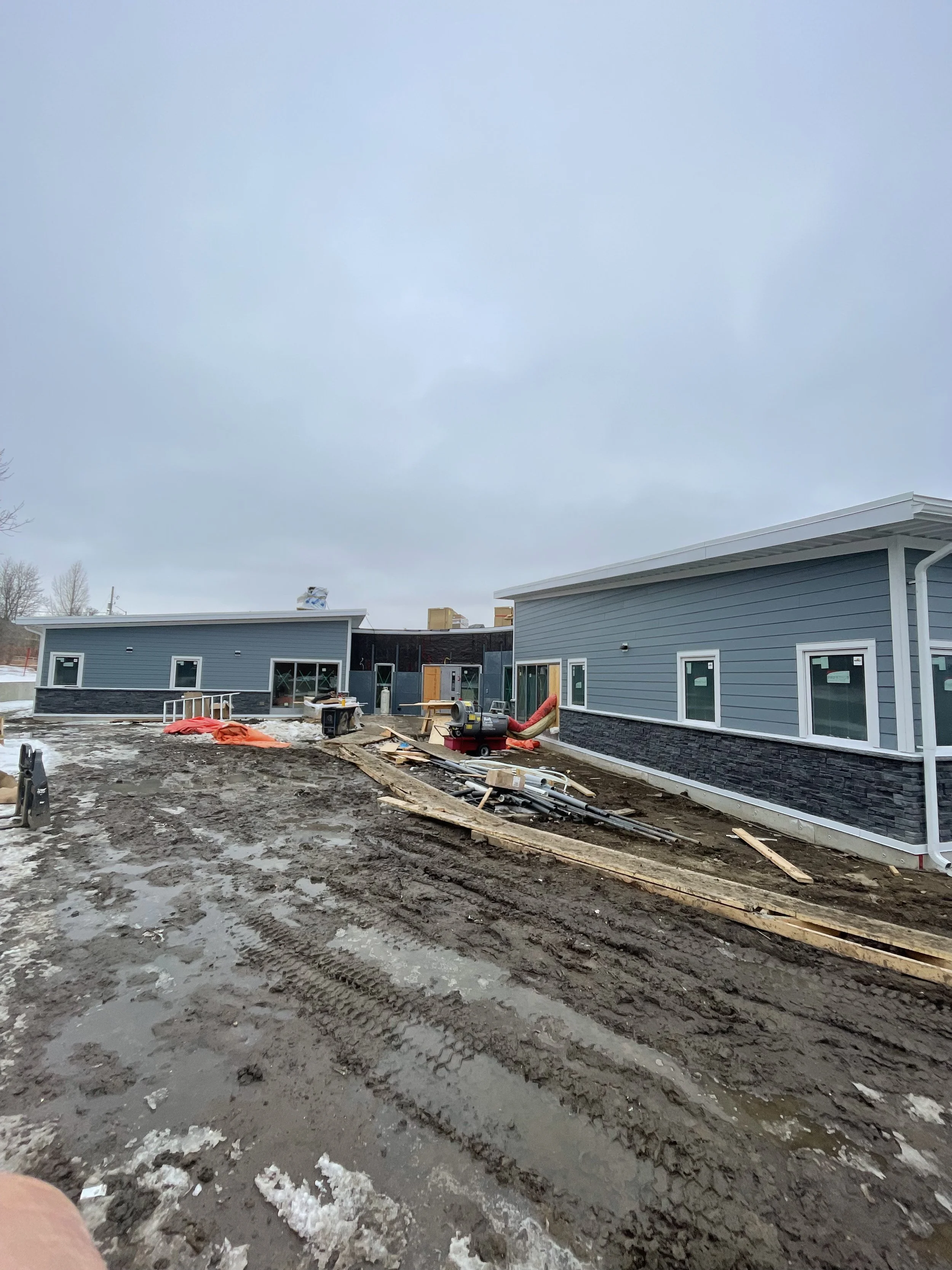 Construction site of a new building with gray siding and white trim, muddy ground, construction materials, and equipment