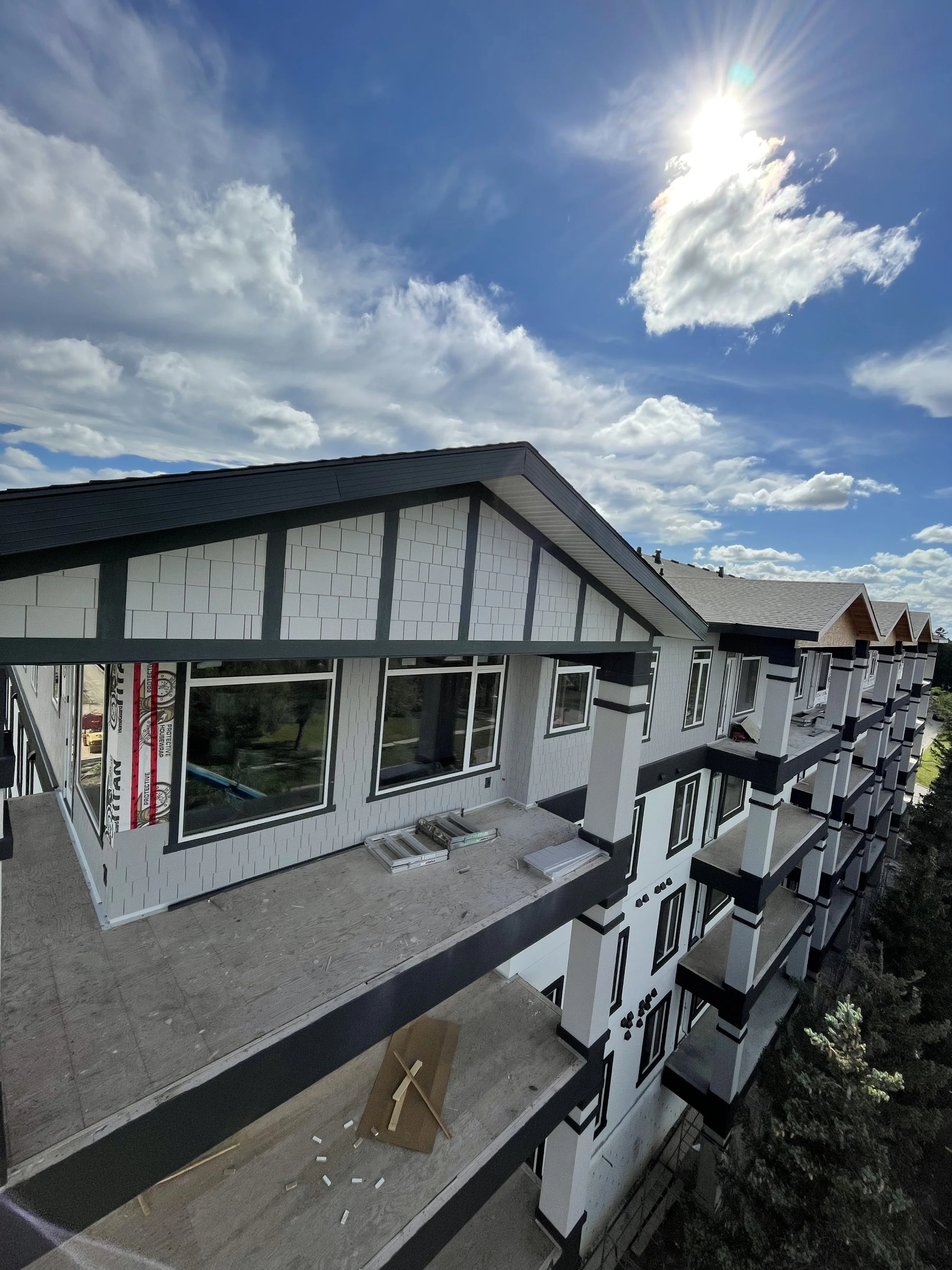 Construction site of a multi-story residential building with some unfinished balconies and large windows, under a partly cloudy sky with the sun shining.