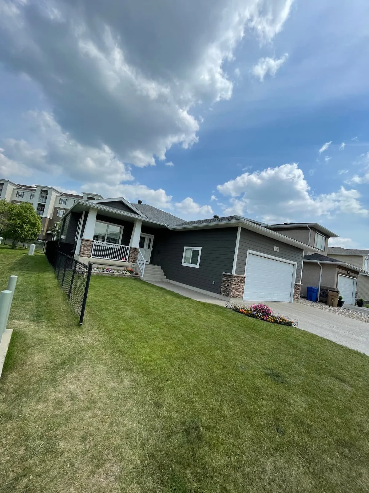 A modern house with a gray exterior, white garage door, and a small covered porch, surrounded by a well-maintained lawn with flower beds and a black fence, under a partly cloudy sky.